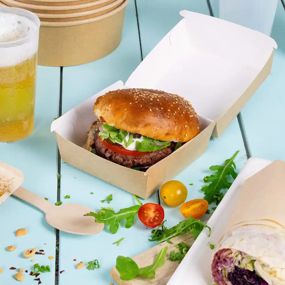 Un burger avec un pain au sésame, de la laitue, une tomate et de la sauce dans une boîte à emporter brune, accompagné de légumes et de bière.