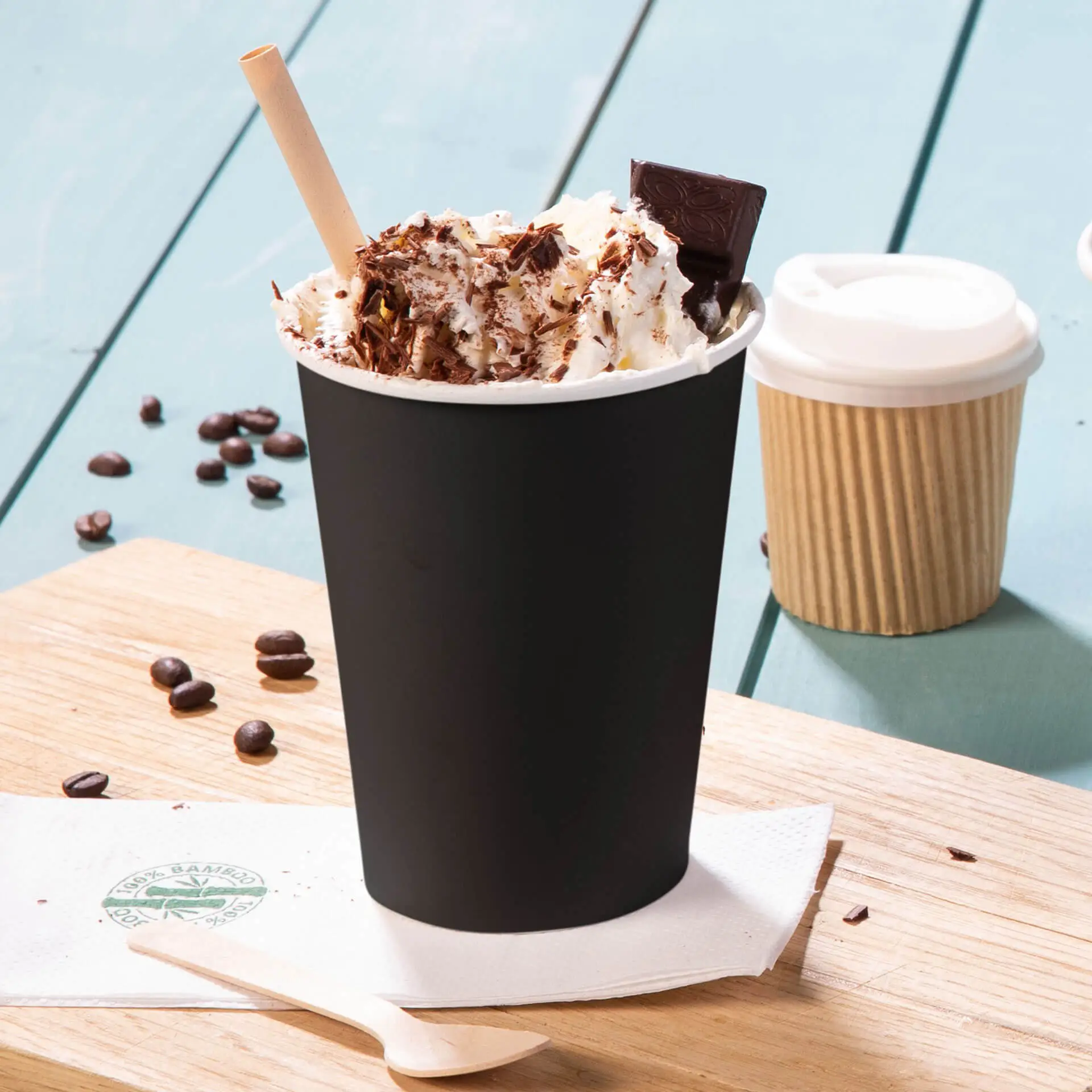 Gobelet en papier noir à paroi unique, garni de crème fouettée, copeaux de chocolat et une paille, servi sur une planche en bois, avec des grains de café à proximité.