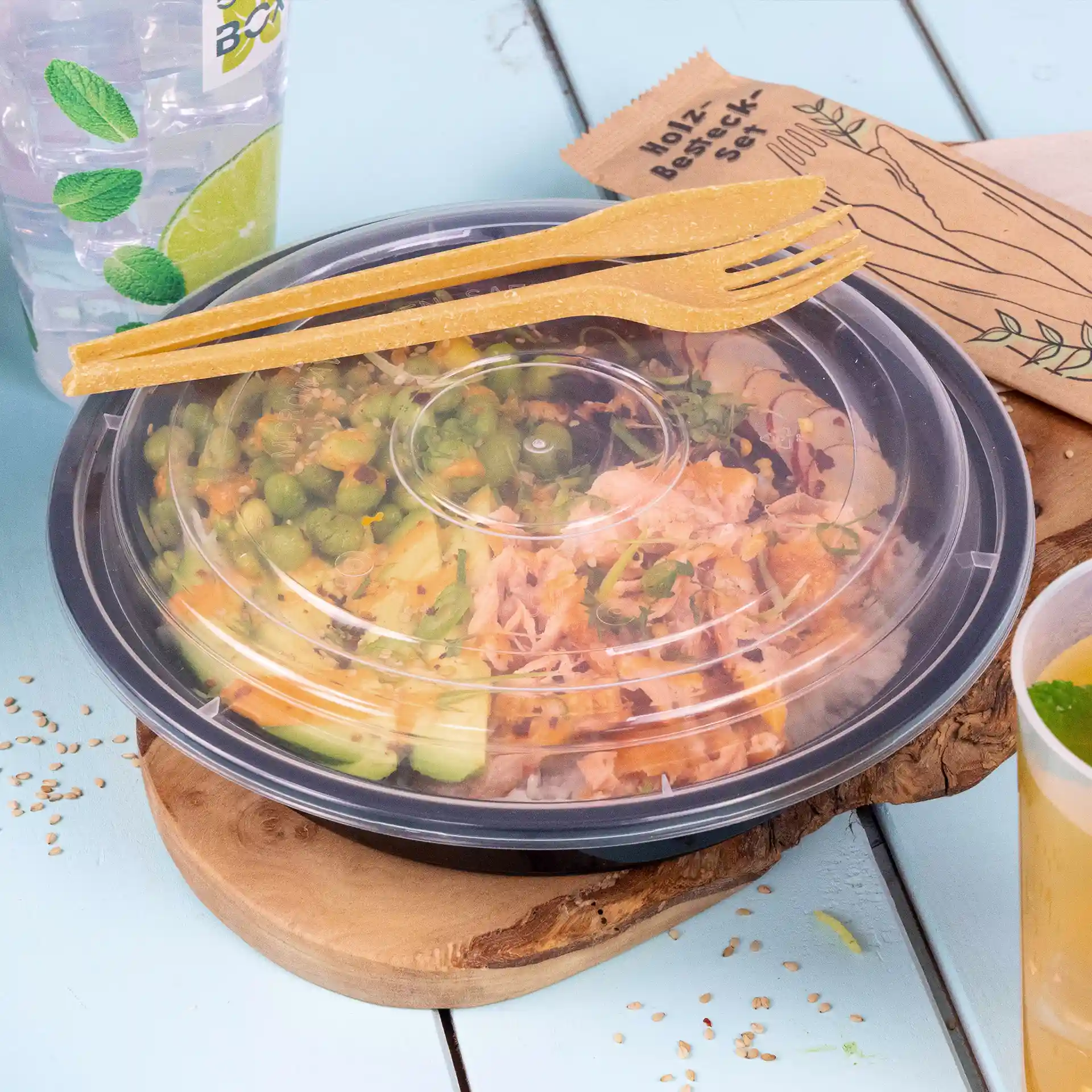 Conteneur avec salade, couvercle transparent et deux fourchettes réutilisables brunes sur une planche en bois, avec une boisson et une serviette à proximité.