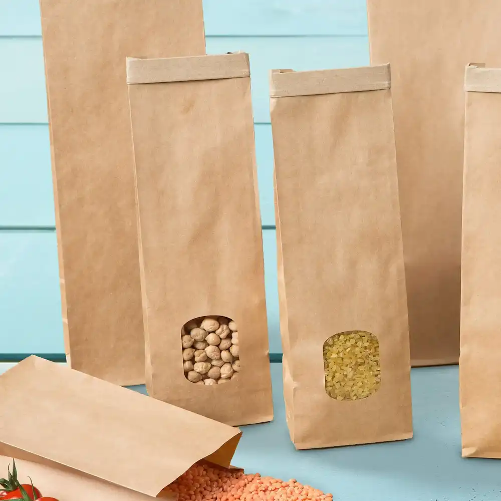 Sacs à fond plat marron avec fenêtres transparentes affichant le contenu, placés sur une surface bleu clair.