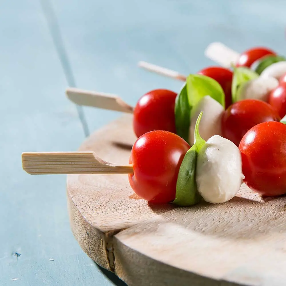 Brochettes de tomates cerises, feuilles de basilic et billes de mozzarella sur une planche en bois.