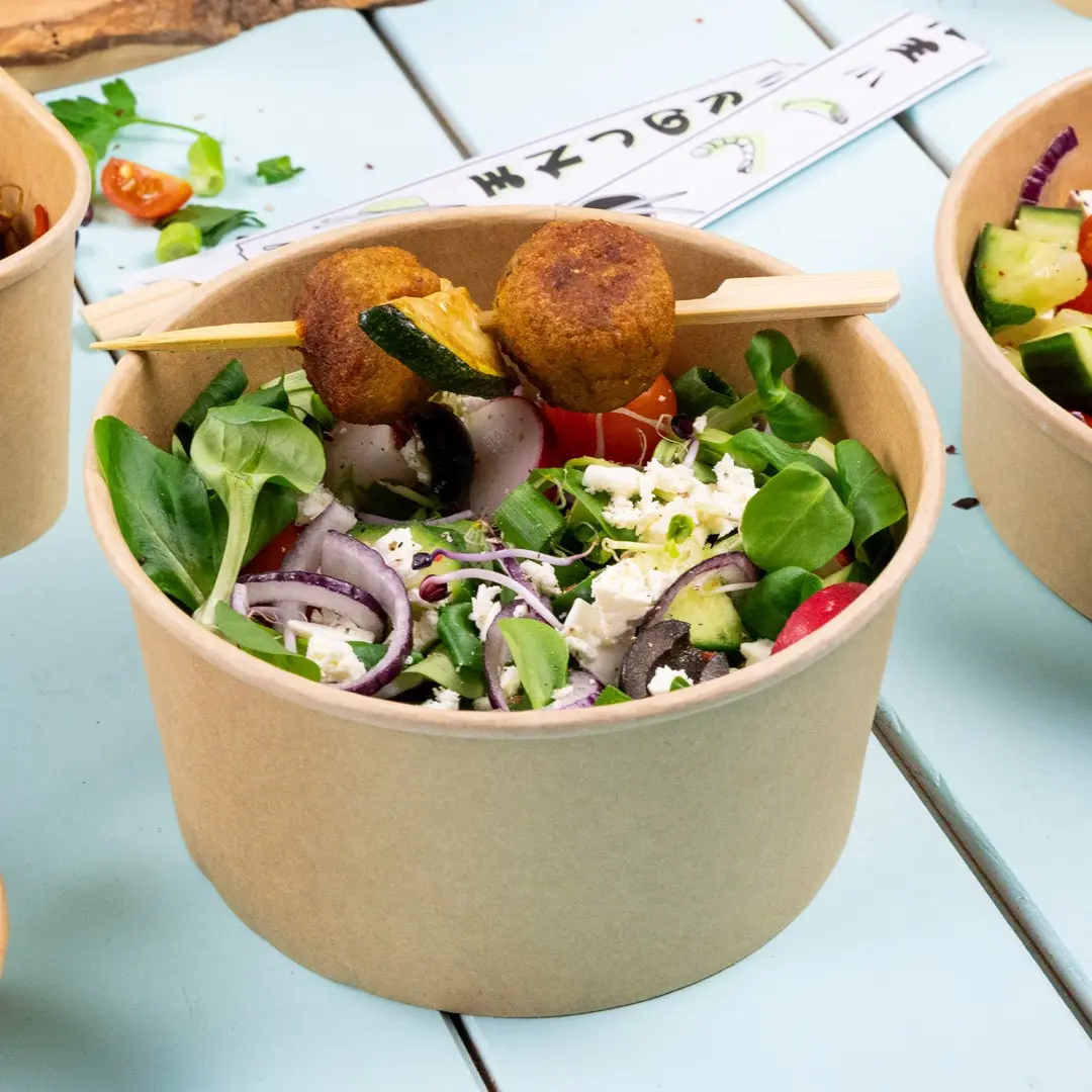 Salade dans un bol en papier brun avec des légumes, tomates, oignons, olives, feta et deux falafels sur une brochette.