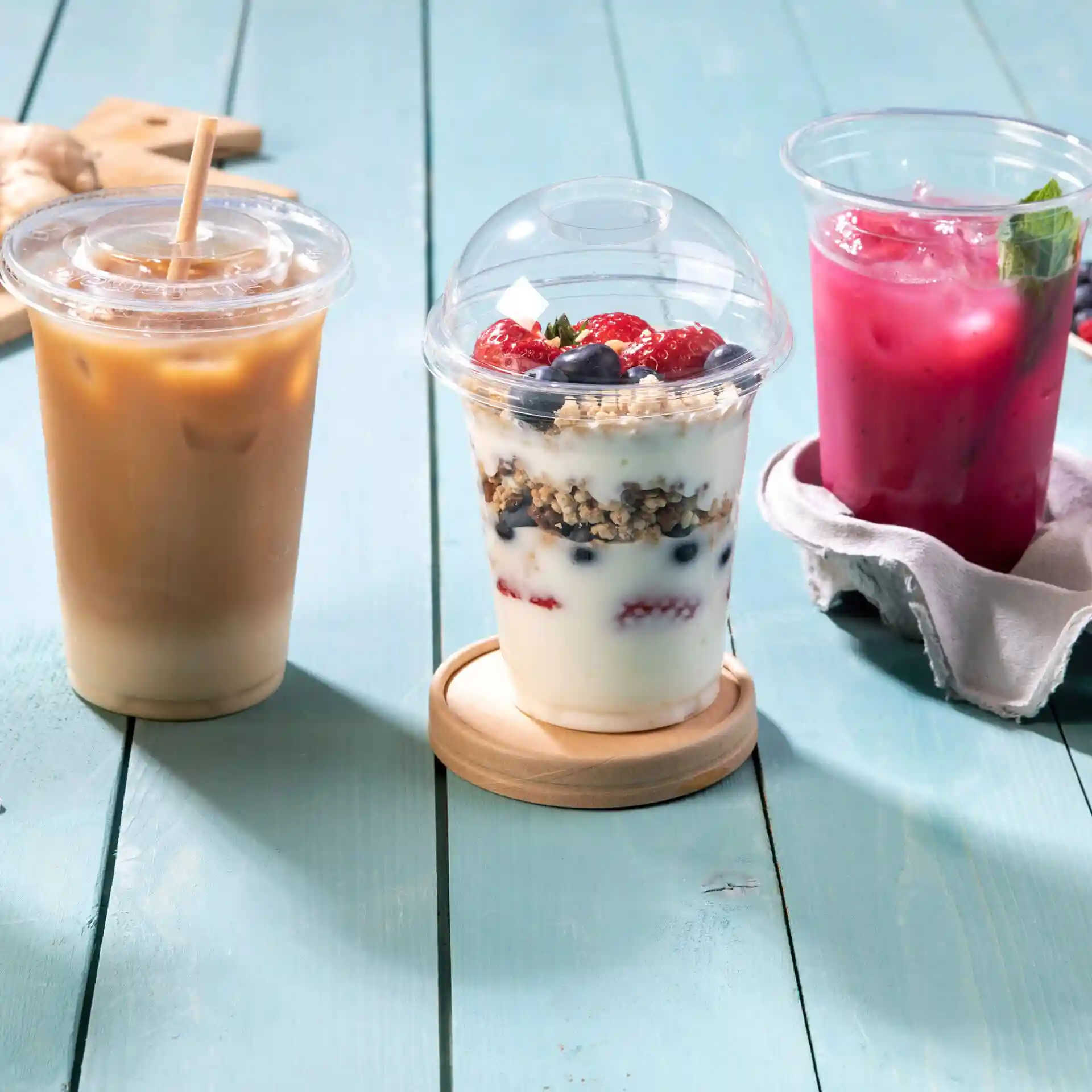 Image de trois boissons : un café glacé, un parfait au yaourt avec des baies et une boisson rose, tous dans des gobelets transparents sur une surface en bois bleu.