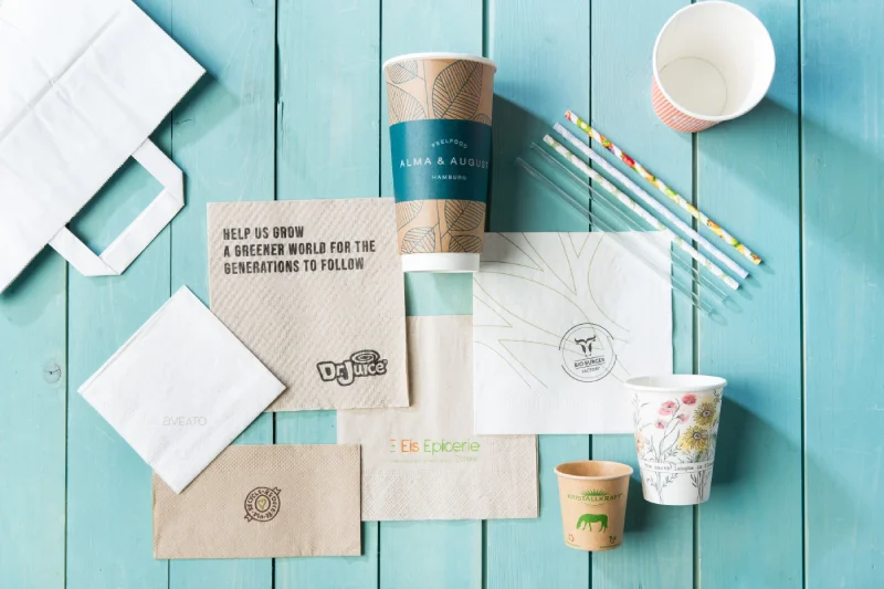 Divers articles d'emballage, y compris des gobelets en papier, des serviettes et des pailles sur une surface en bois turquoise.