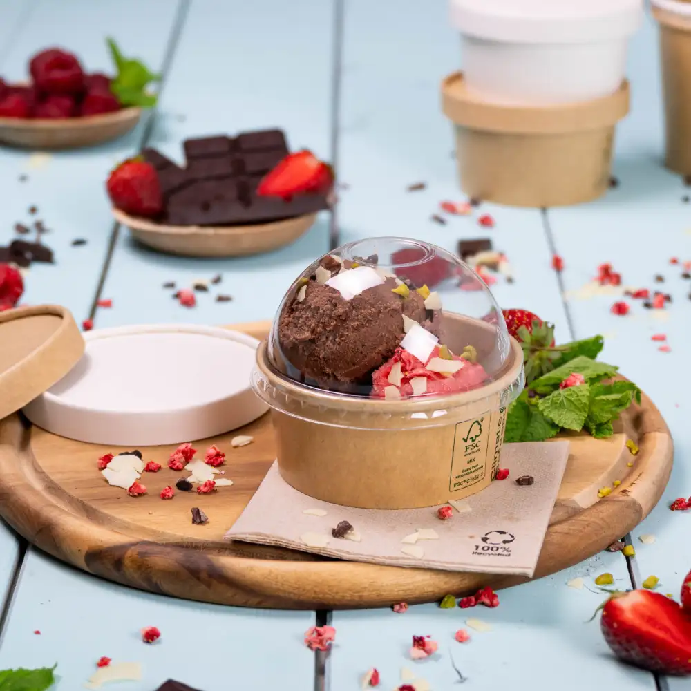 Crème glacée dans un contenant en dôme transparent sur un plateau en bois, garnie de fraises, de menthe et de morceaux de chocolat.