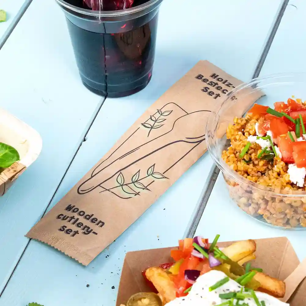 Emballage d'un ensemble de couverts en bois à côté d'un gobelet en plastique, d'un bol de salade de céréales et d'un contenant en carton de frites sur une surface bleue.