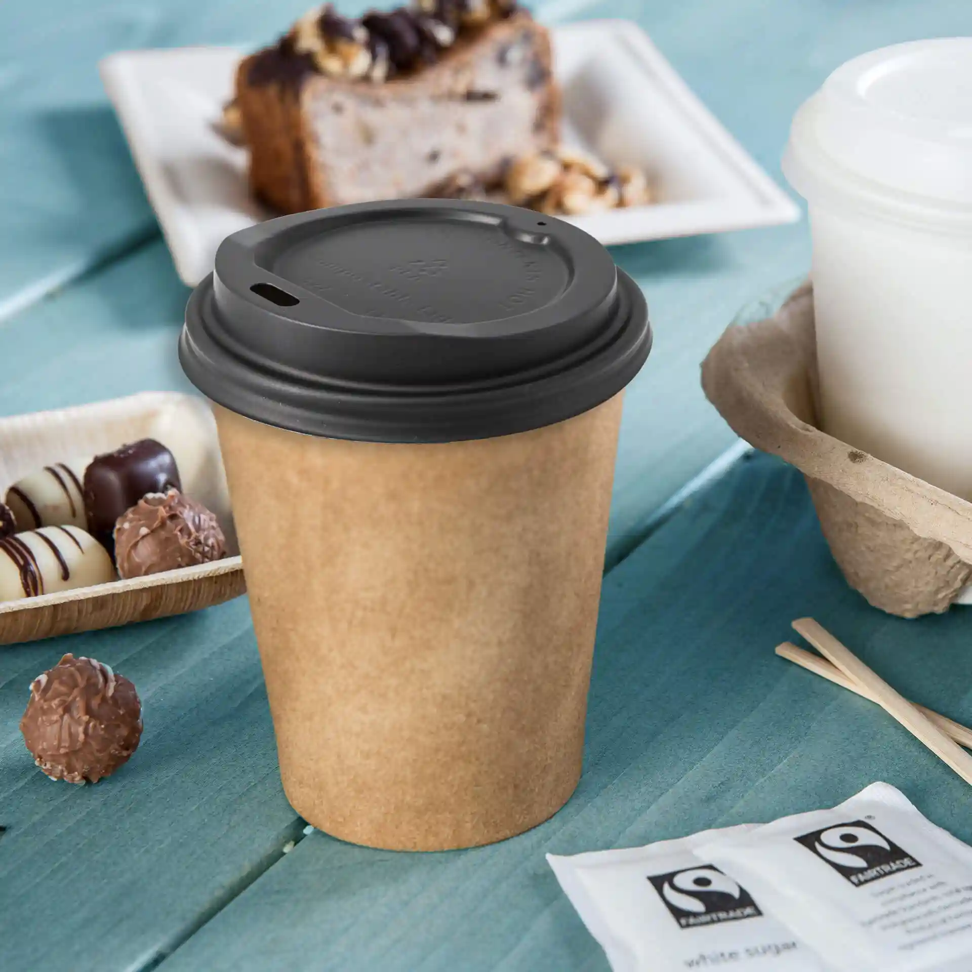 Couvercle de tasse en CPLA noir avec un trou pour boire, posé sur une surface en bois bleu avec des collations à proximité.