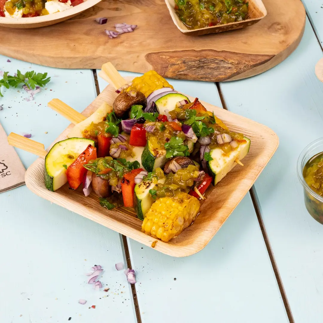 Brochettes en bois avec des légumes grillés, y compris courgette, poivron, maïs et champignons, garnis de sauce dans un plateau en bois.