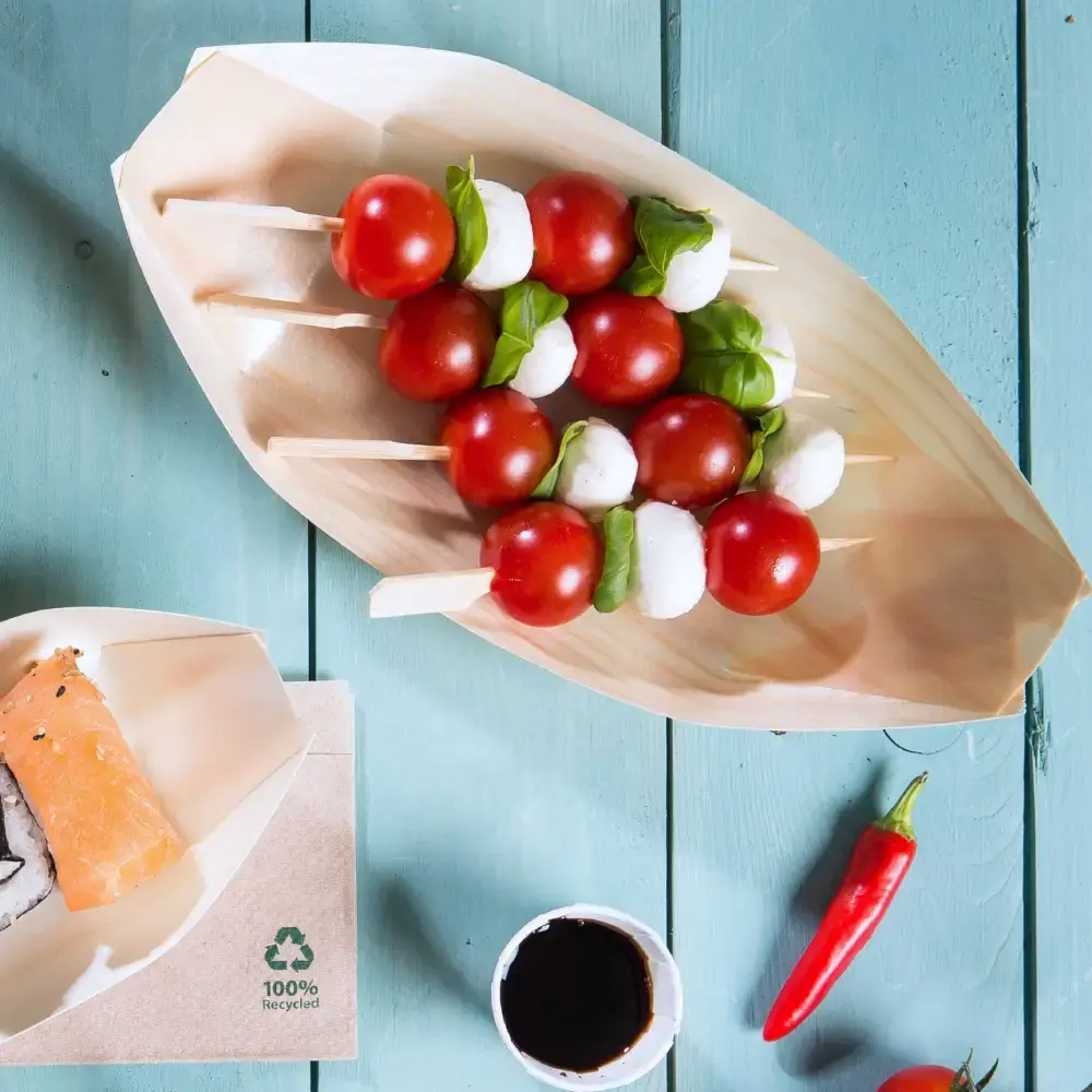 Plateau en bois en forme de bateau avec des brochettes de tomates cerises, billes de mozzarella et feuilles de basilic, accompagné de sushi et de sauce à tremper.
