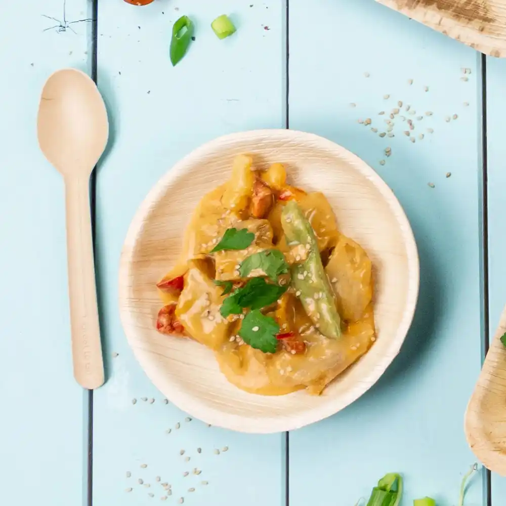 Bol en bambou avec légumes sautés et sauce, garni de graines de sésame et de coriandre, à côté d'une cuillère en bois.
