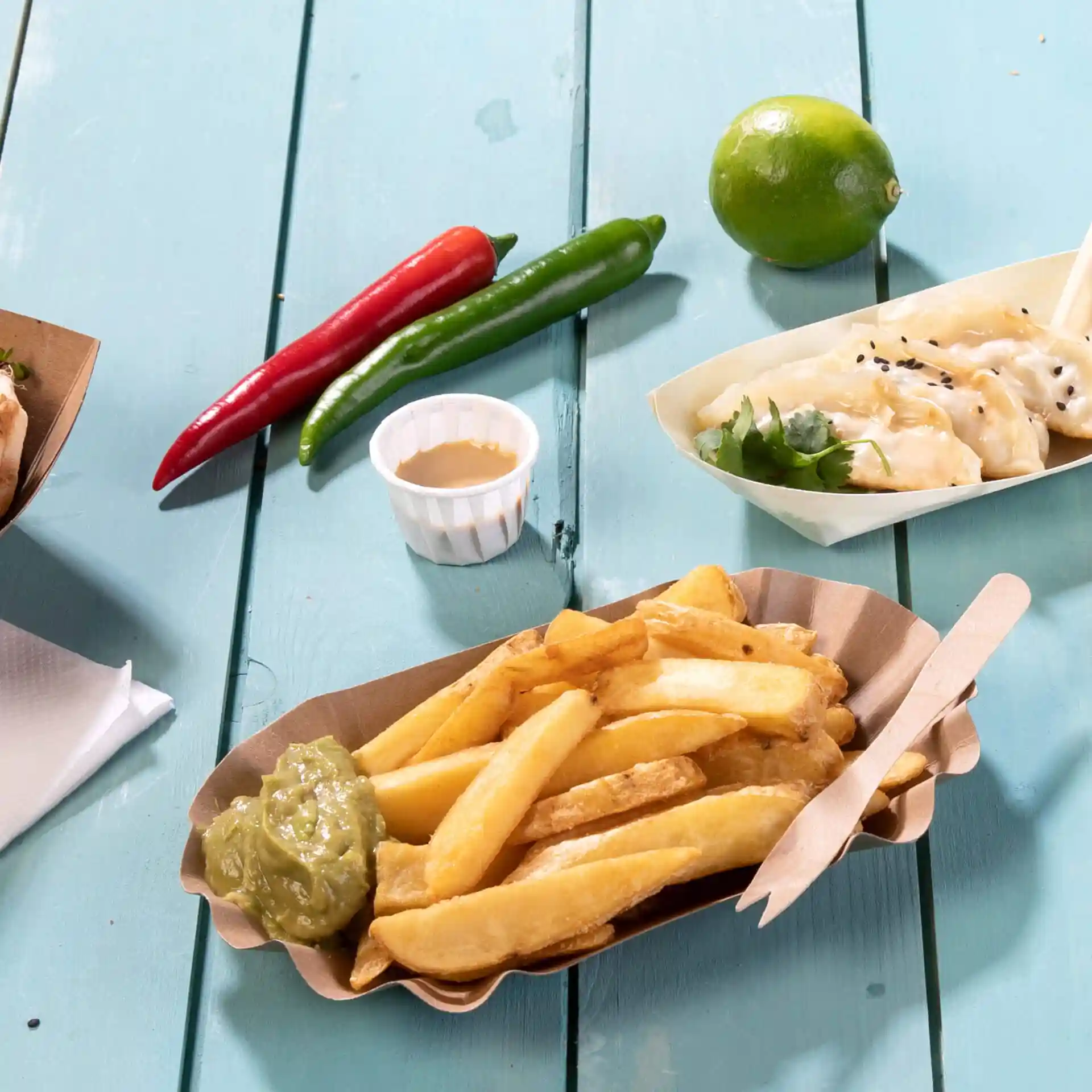 Image d'une portion de frites avec une sauce verte dans un plateau en papier, entourée de piments, d'un citron vert et d'un petit pot de sauce.