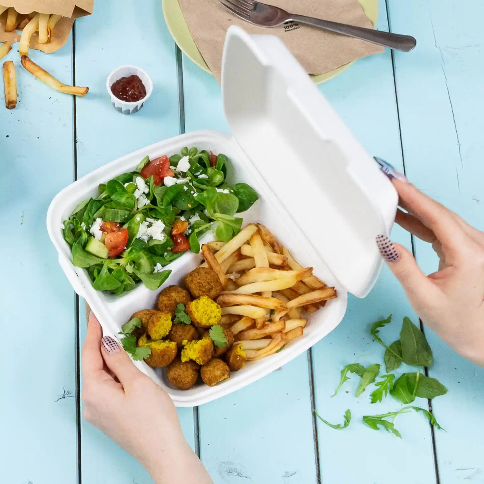 Une main tenant un contenant blanc à couvercle articulé avec deux compartiments, rempli de salade, frites et boulettes de falafel.