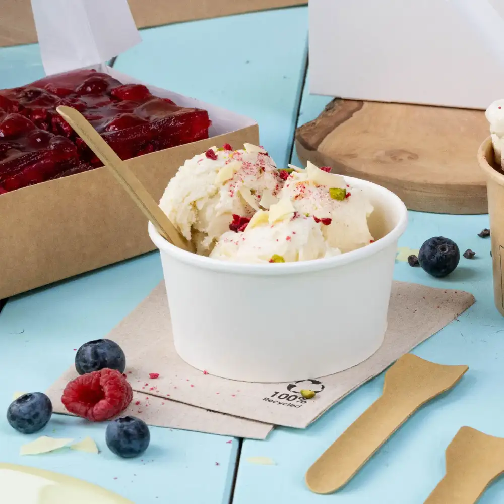 Glace dans un verre blanc, garnie de noix et de baies, à côté d'une boîte à dessert, avec des cuillères en bois et des fruits frais sur une surface bleue.
