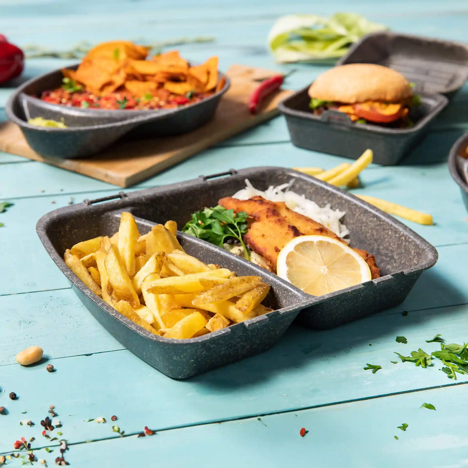 Conteneur de repas réutilisable gris avec deux compartiments, rempli de frites et de poisson, garni de citron et d'herbes.