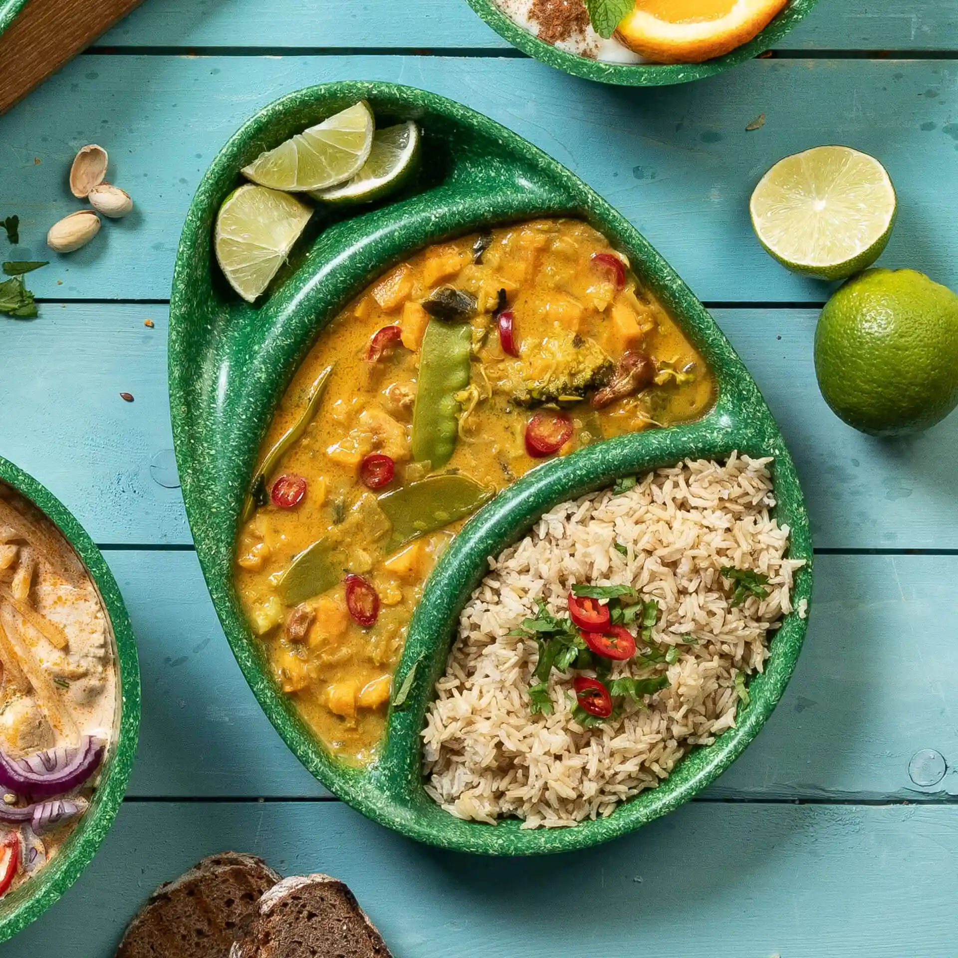 Assiette réutilisable verte à trois compartiments, remplie de curry, de riz et de quartiers de citron vert sur une surface en bois bleu.