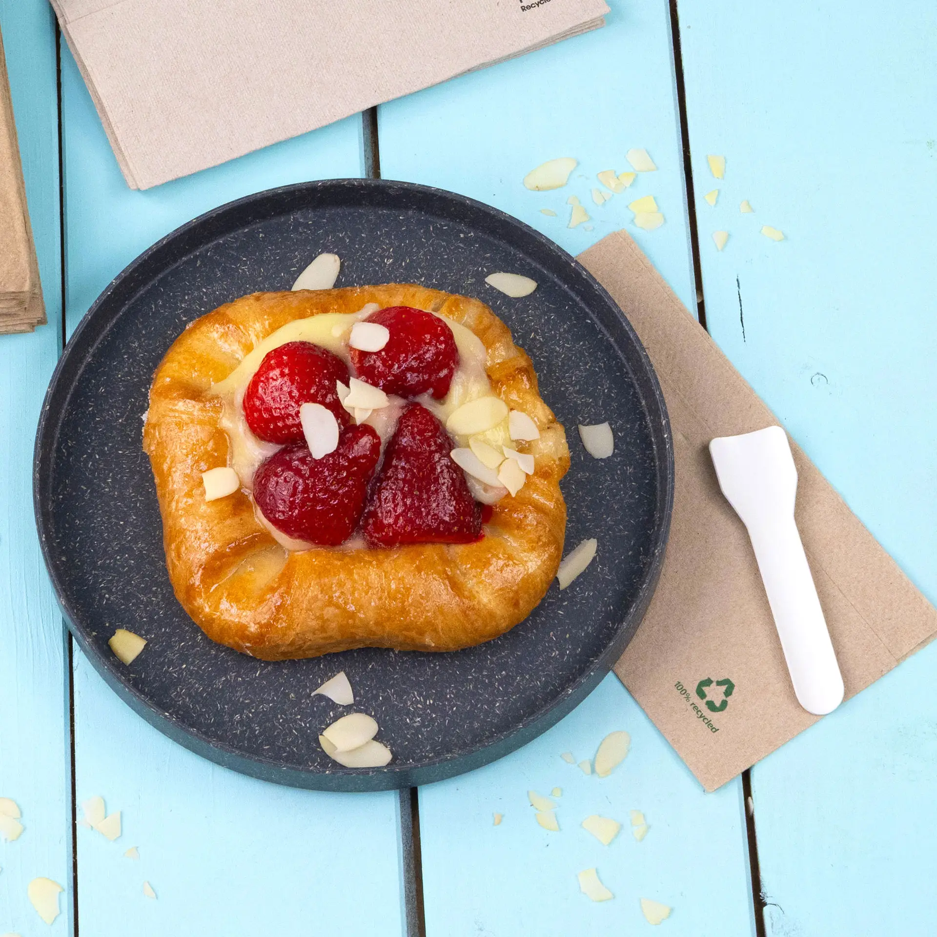 Pâtisserie avec des fraises et des amandes en tranches sur une assiette noire, accompagnée d'un ustensile en plastique blanc et d'une serviette non blanchie.