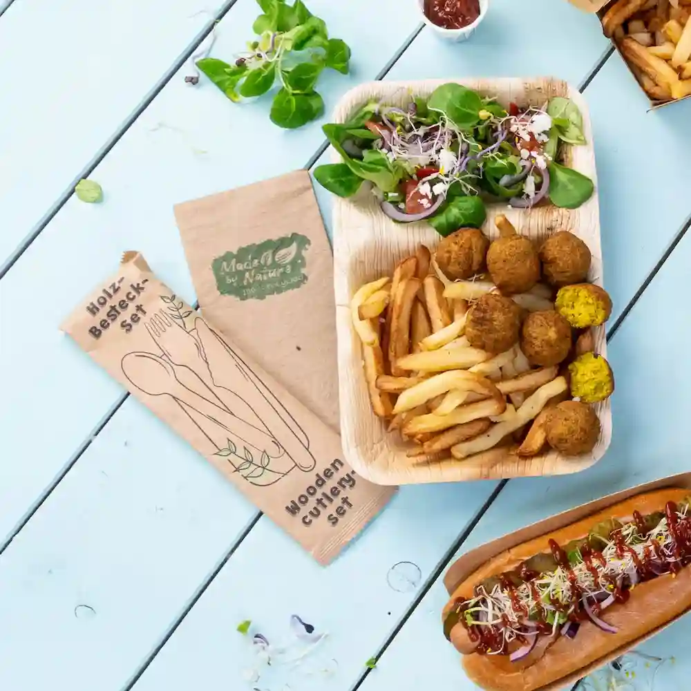 Ensemble de couverts en bois à côté d'un plateau de nourriture avec salade, frites, boulettes de falafel et un hot-dog sur une table bleue.