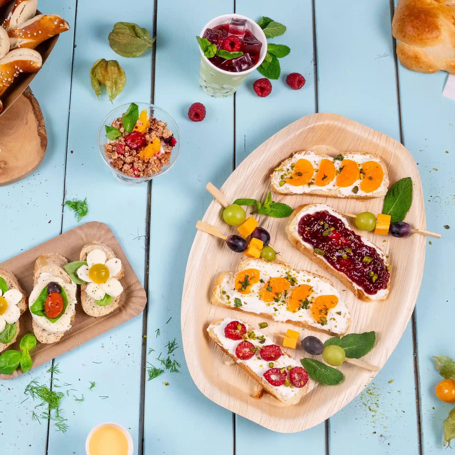 Plateau rectangulaire en feuille de palmier avec des sandwiches ouverts variés, garnis de fruits et d'herbes, sur une table en bois bleu.