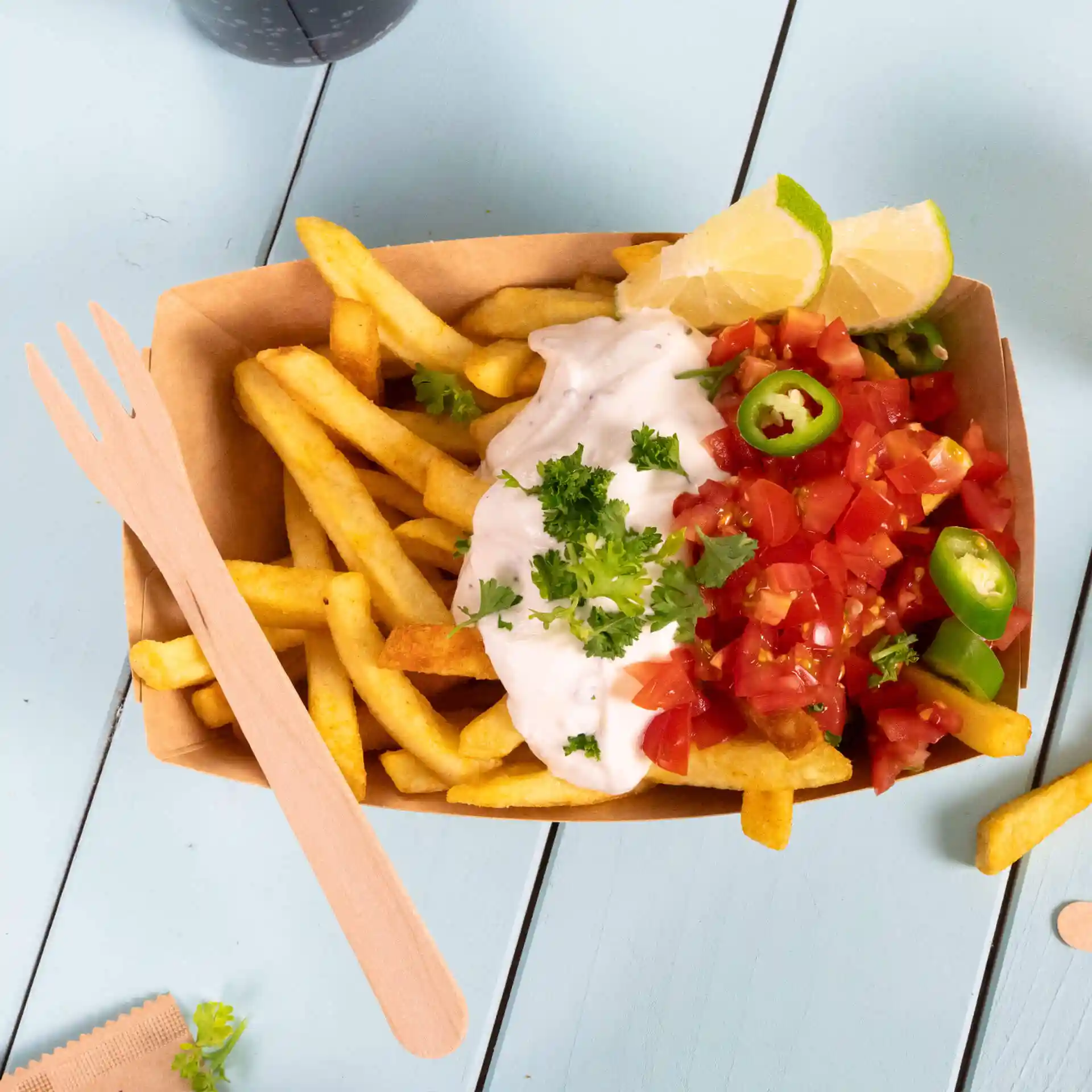 Fourchette en bois à côté d'une portion de frites garnie de crème aigre, de tomates en dés, de poivrons verts et de quartiers de citron vert.