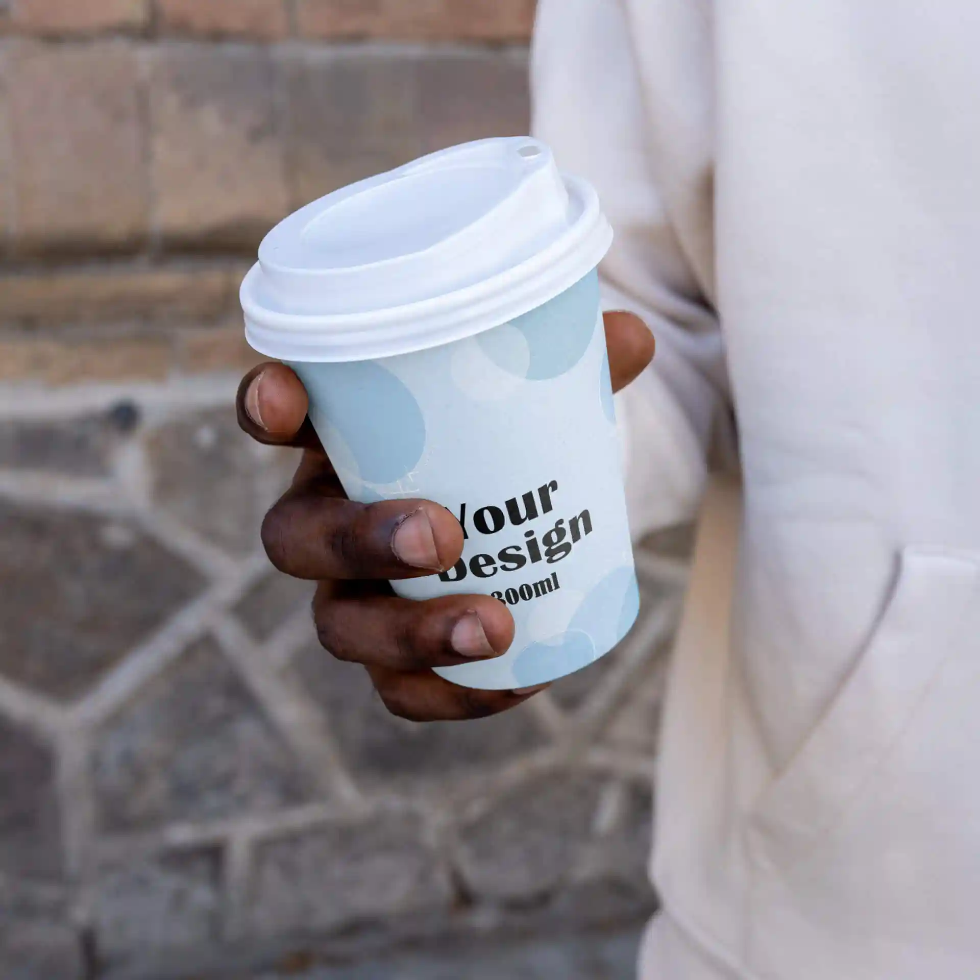 Une main tenant un gobelet en papier blanc avec un design bleu clair et un couvercle blanc, portant le texte "Votre design" et un volume de 300 ml.