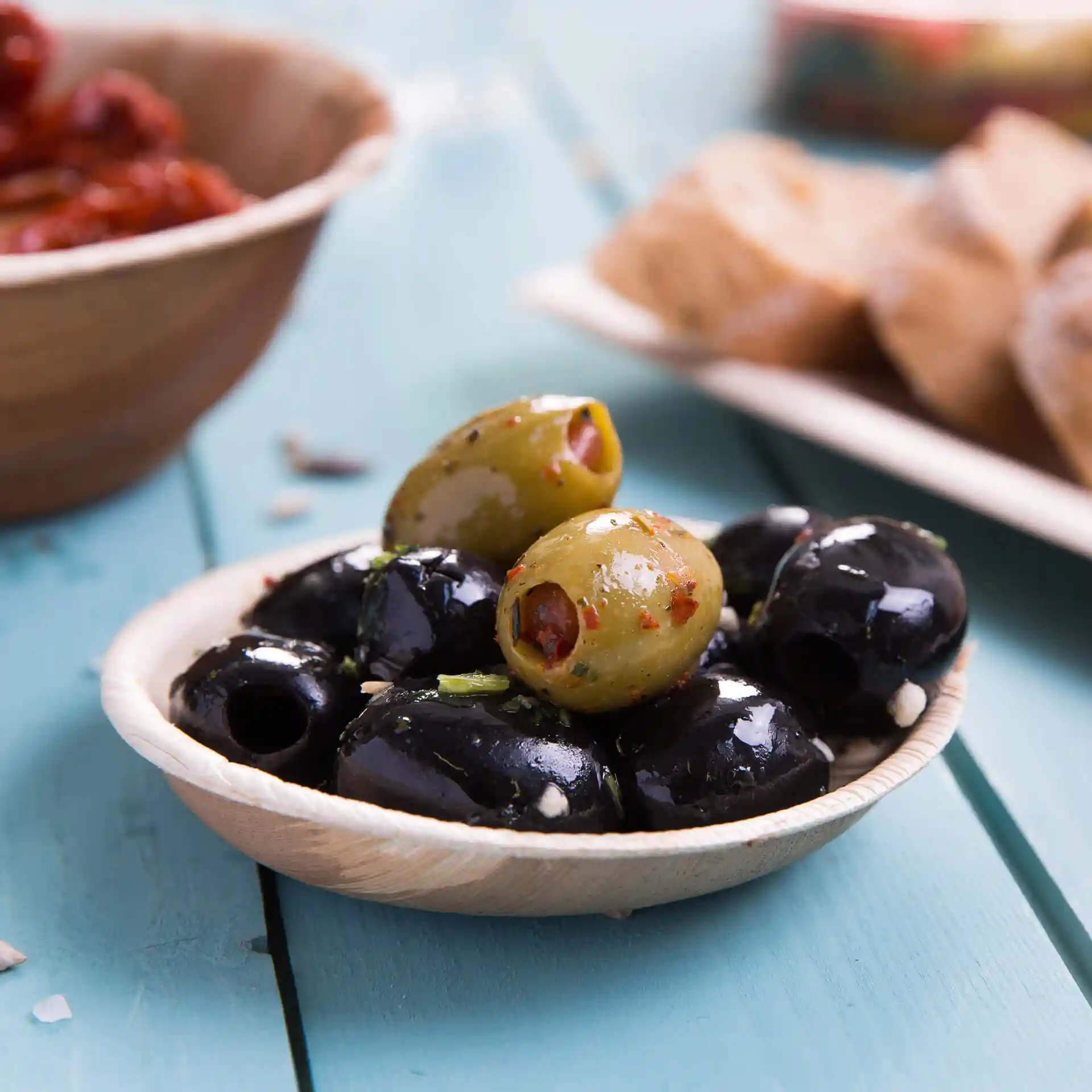 Bol en feuille de palmier contenant des olives noires et vertes, posé sur une surface en bois bleu.