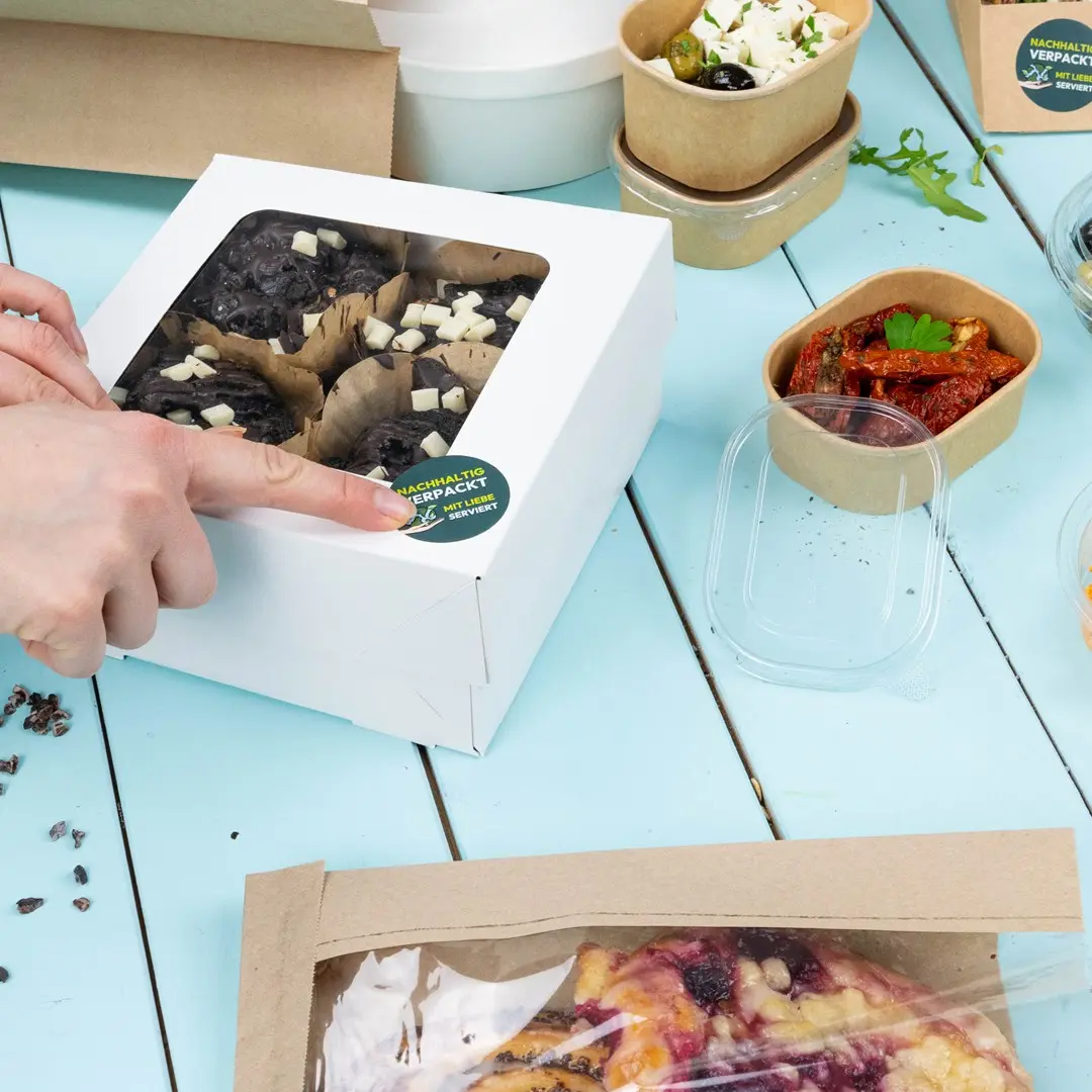 Boîte blanche contenant des produits de boulangerie avec un couvercle transparent, entourée de divers contenants alimentaires sur une table bleue.