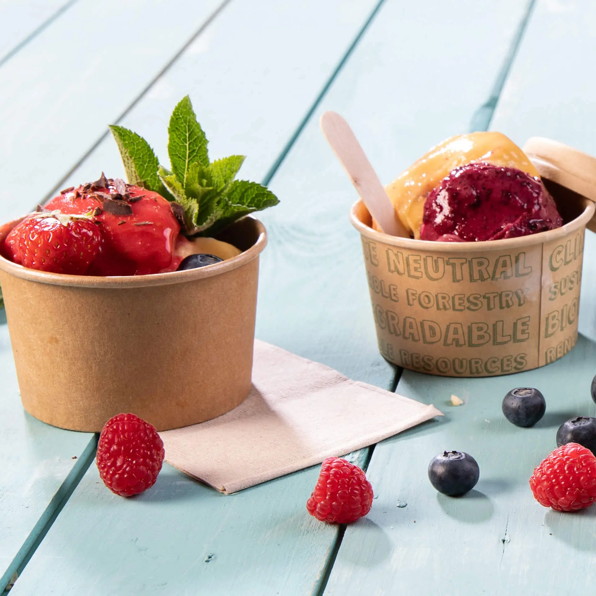 Deux gobelets en papier avec de la glace et des garnitures de fruits, l'un avec des fraises et de la menthe, l'autre avec de la glace violette et jaune.