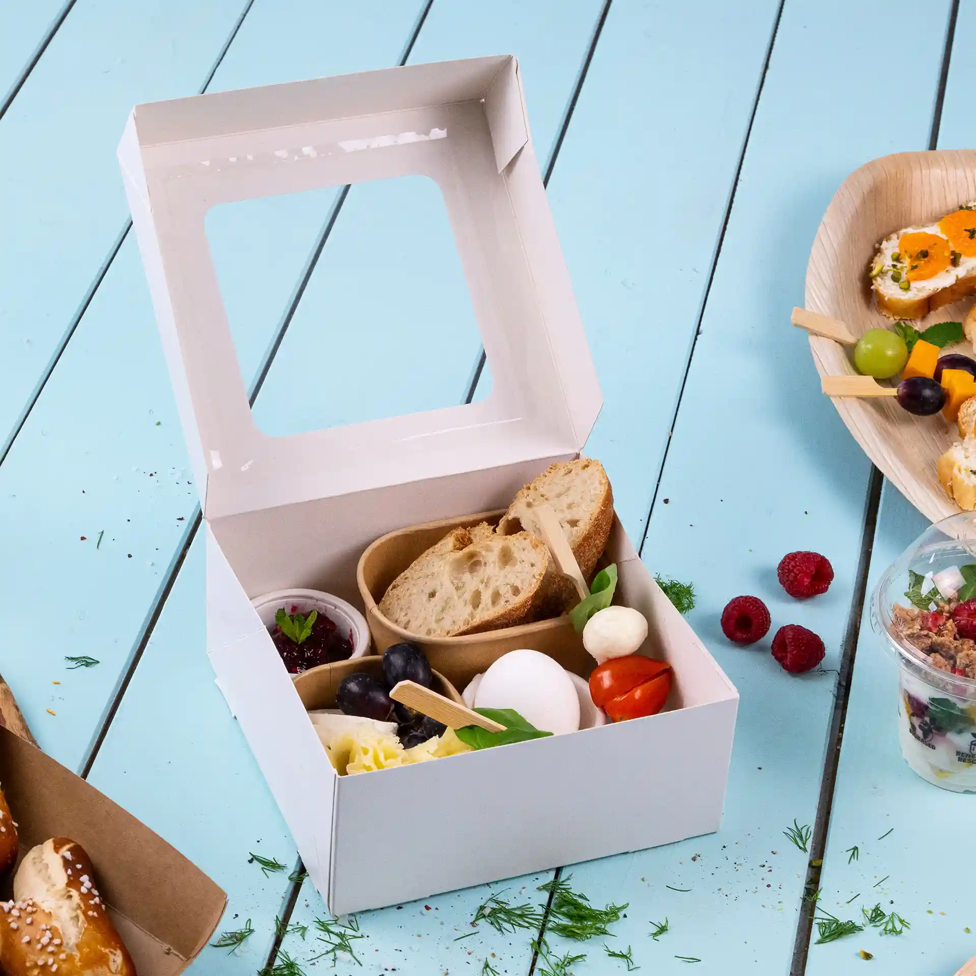 Boîte en carton blanc avec une fenêtre en cellulose, contenant du pain, du fromage et des fruits assortis.