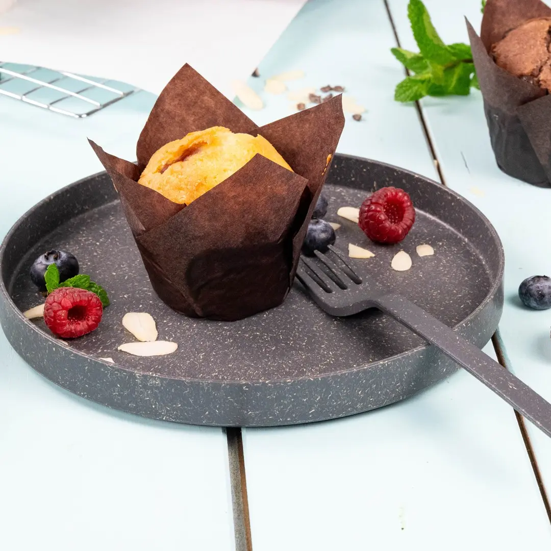 Muffin dans un emballage en papier brun sur une assiette grise avec des framboises, des myrtilles, des amandes en tranches et une petite fourchette.