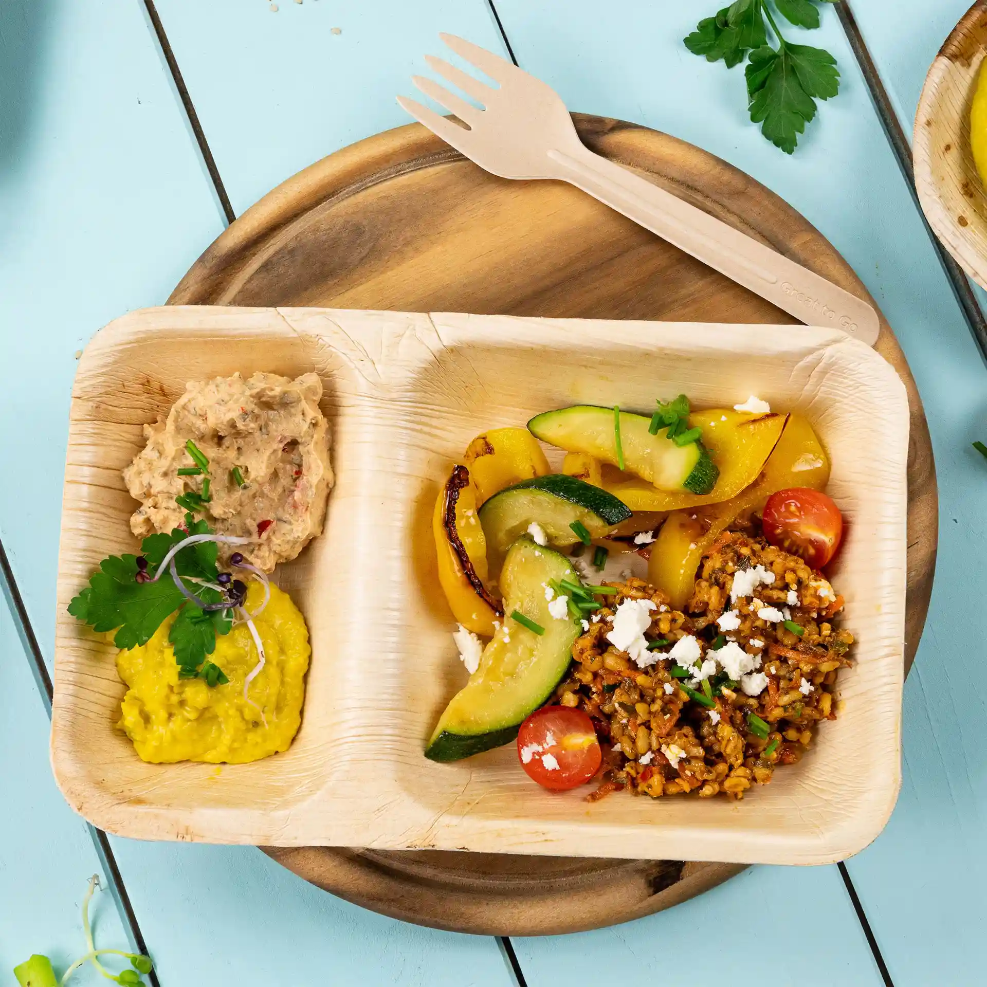 Assiette rectangulaire en feuille de palmier avec deux compartiments, remplie de légumes, de céréales et de dips, sur une surface en bois.