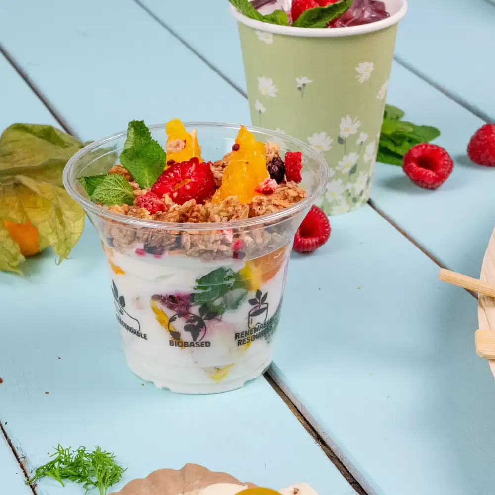 Gobelet à dessert en plastique transparent rempli de yaourt, de fruits et de granola, placé sur une surface bleue avec des feuilles de menthe à proximité.
