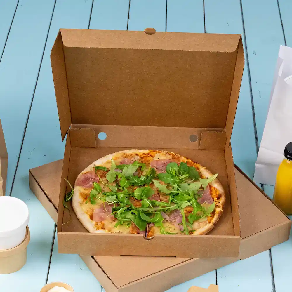 Pizza dans une boîte à emporter en carton, garnie de jambon et de mélanges de légumes sur une surface en bois bleue.