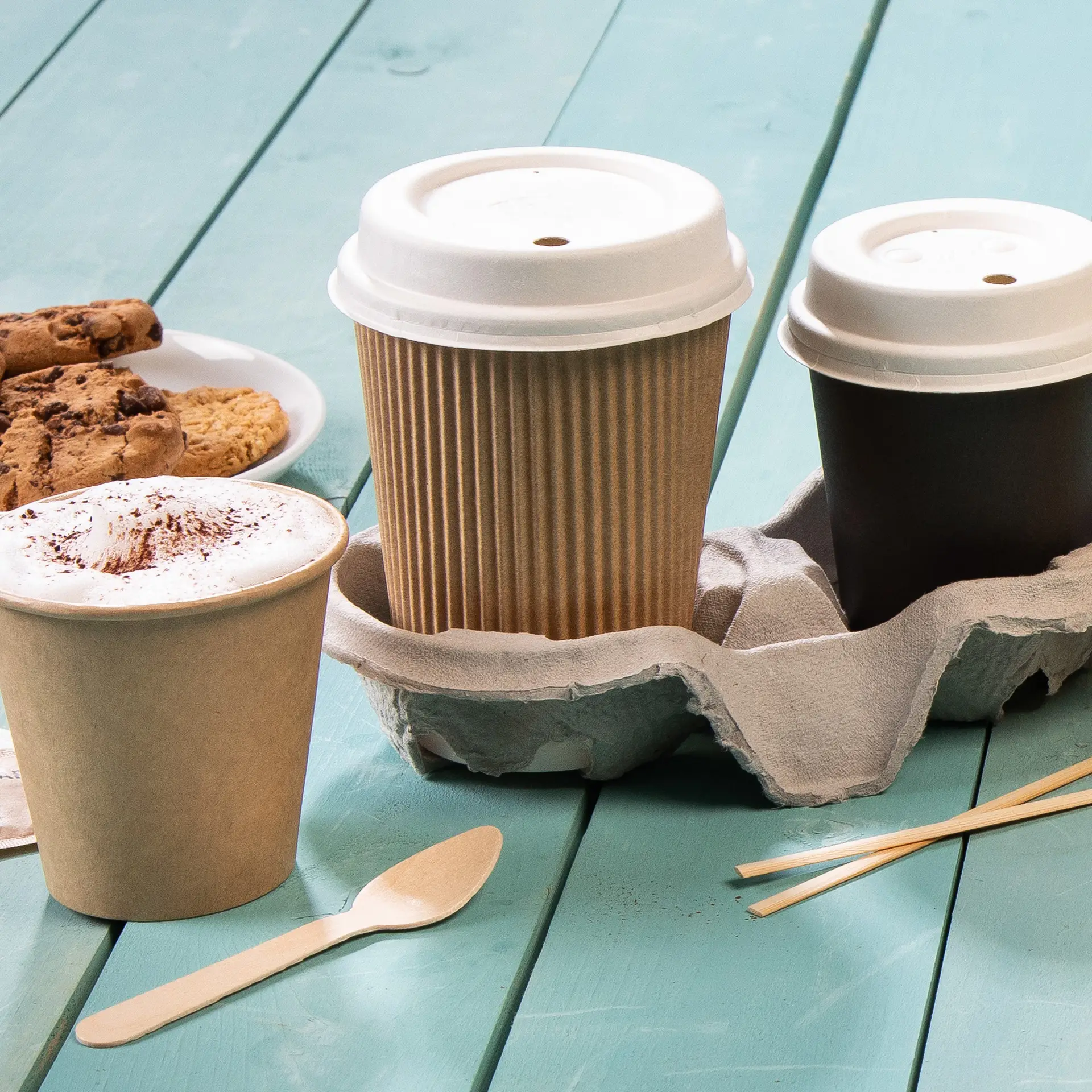 Une tasse à café isotherme marron, un gobelet en papier marron et un gobelet en papier noir dans un porte-gobelet gris, avec des biscuits à côté.