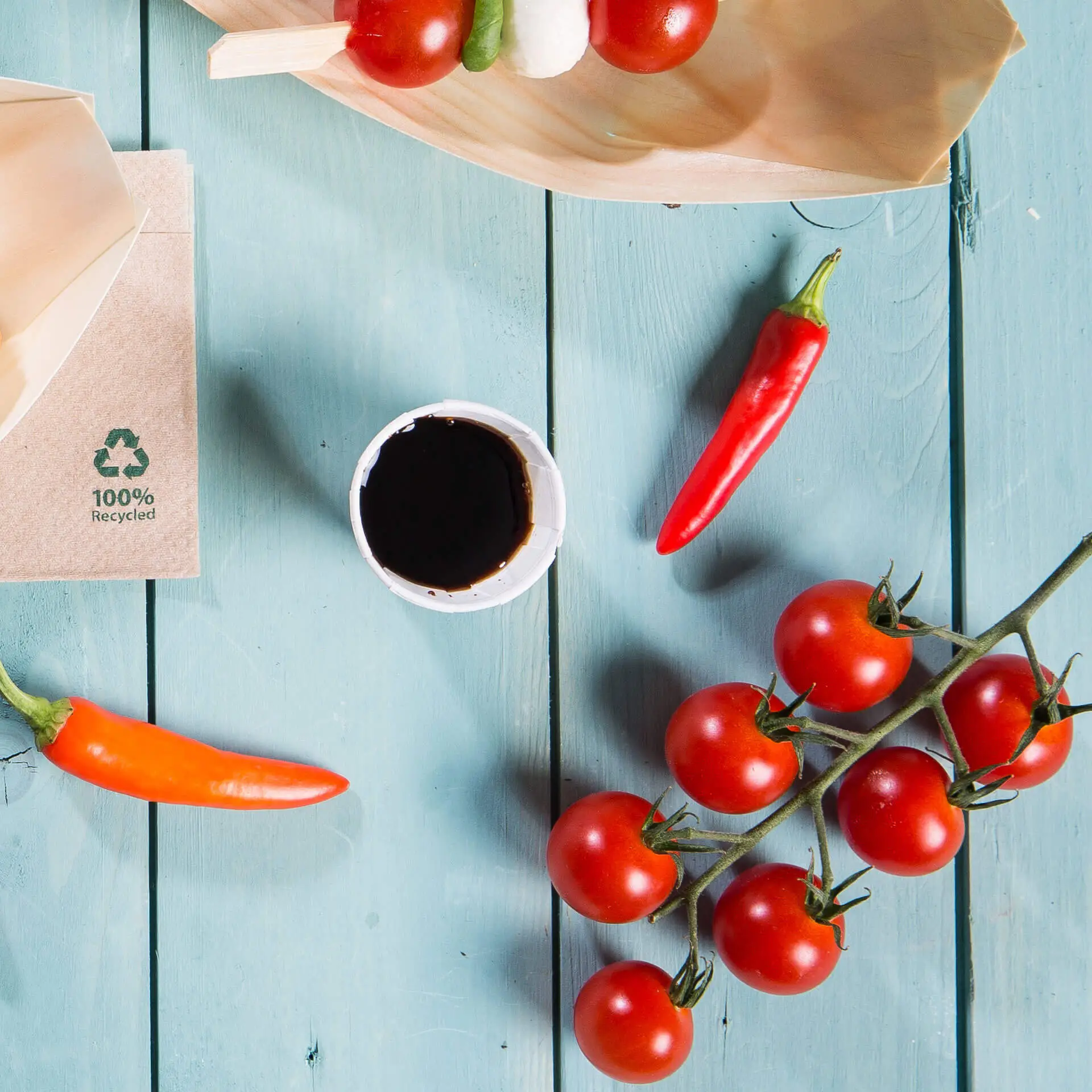 Image d'une assiette en bois avec des tomates cerises, des piments, un petit gobelet en papier blanc avec une sauce foncée et un emballage brun avec un symbole de recyclage.