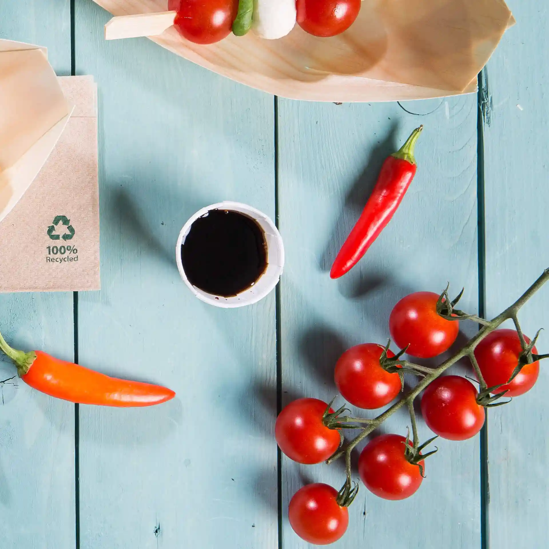Image d'un gobelet en papier avec un liquide sombre, entouré de tomates cerises, de piments et d'une assiette en bois sur une surface en bois bleu.