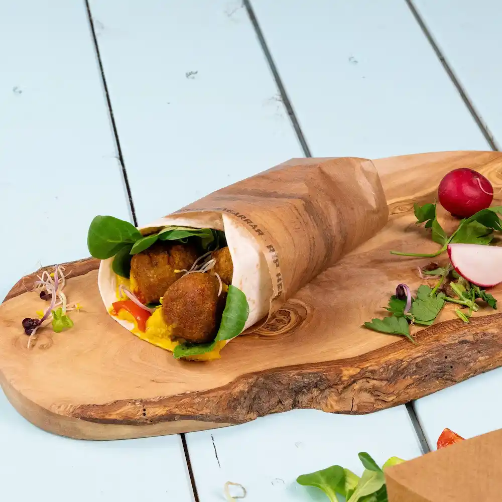 Un en-cas enveloppé rempli de falafel, d'épinards et de sauce sur une planche en bois, accompagné de radis et d'herbes.