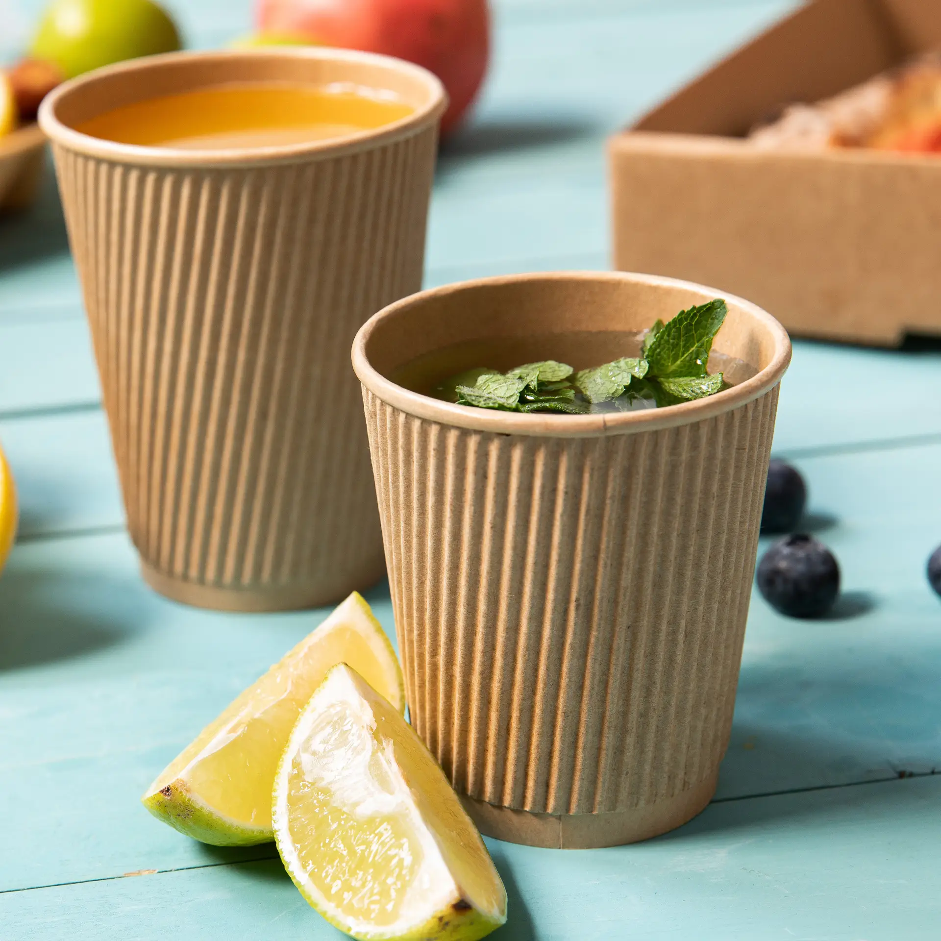 Deux gobelets jetables isothermes marron, l'un avec des feuilles de menthe et des quartiers de citron vert à côté.