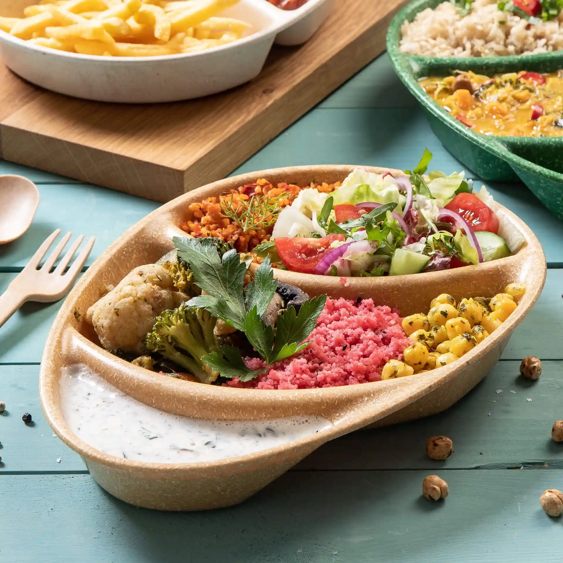 Assiette réutilisable brune à trois compartiments, remplie de divers aliments et d'une sauce sur une surface en bois bleu.