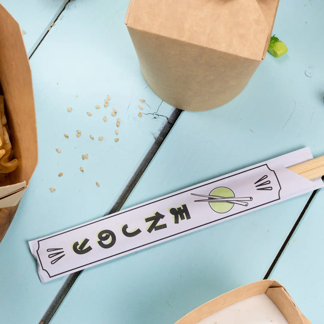 Emballage de baguettes avec design graphique et baguettes sur une surface en bois bleu, entouré de contenants alimentaires et de graines de sésame.