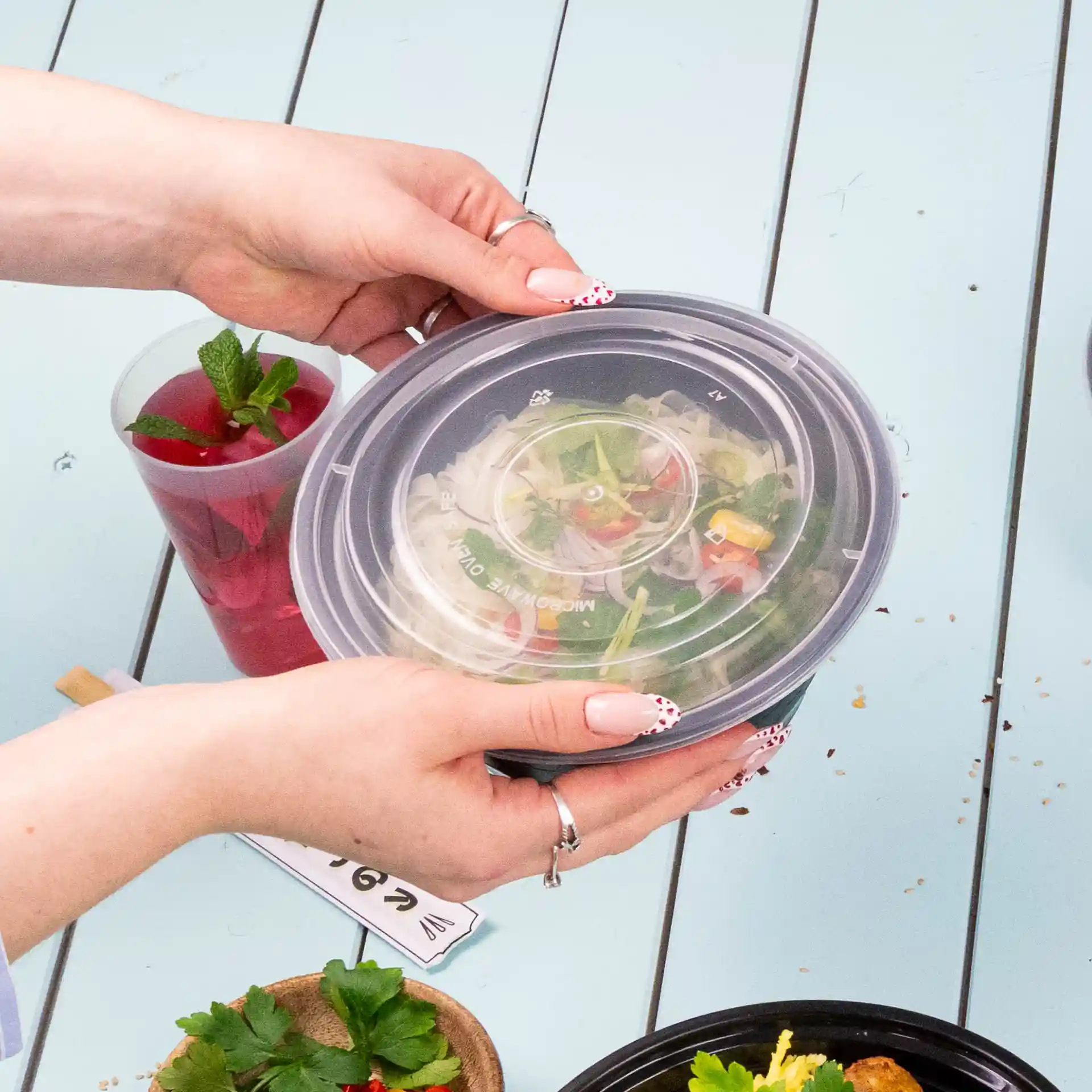 Une personne tient un couvercle rond transparent réutilisable au-dessus d'un bol de salade sur une table bleu clair.