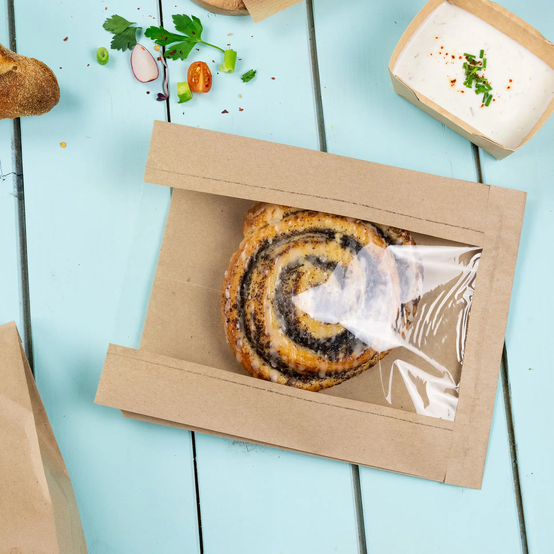 Un sac à pain en papier brun plat avec une fenêtre en PLA transparent montrant une pâtisserie en spirale à l'intérieur ; garnitures et condiments supplémentaires à proximité.