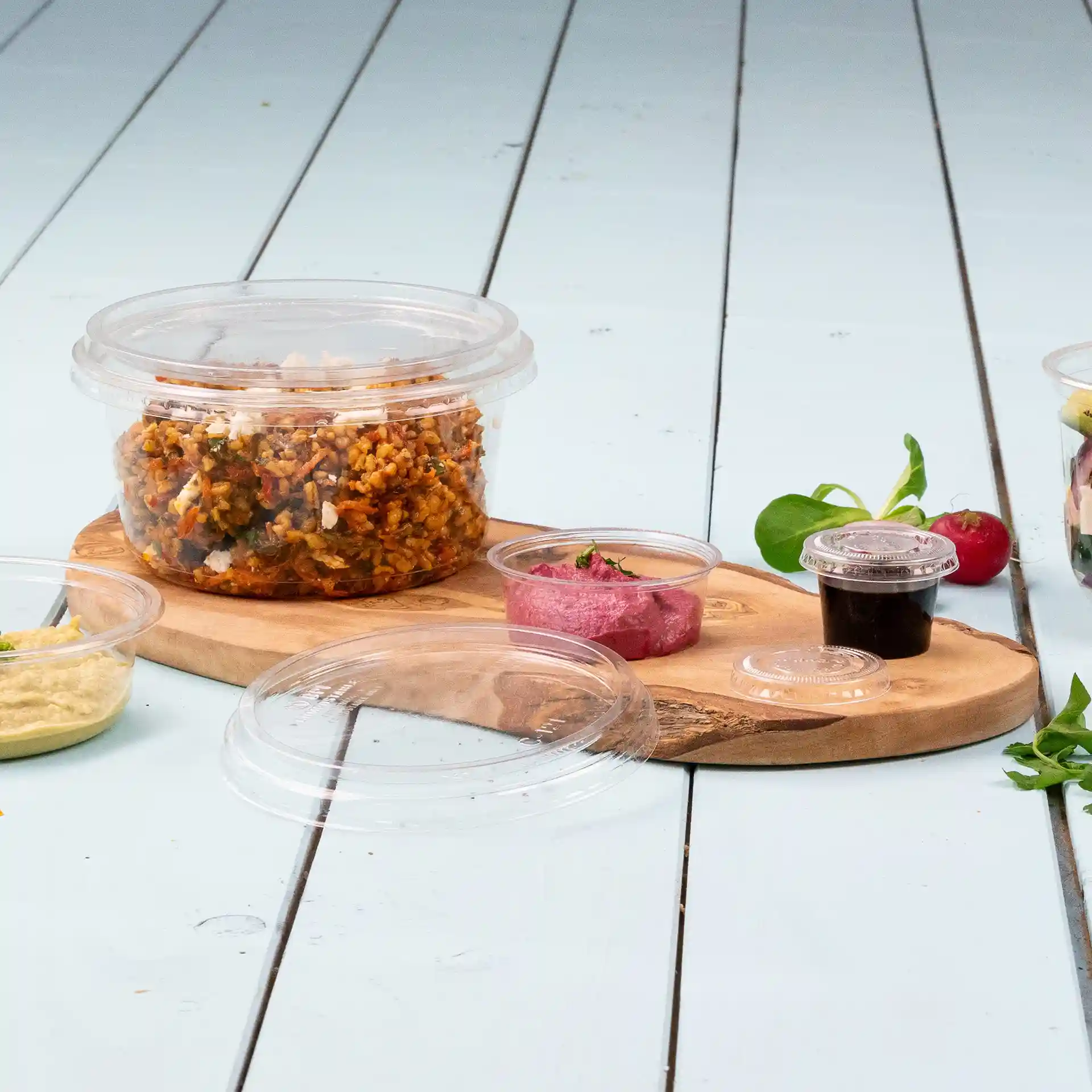 Conteneur rond transparent avec couvercle, rempli de nourriture, sur une planche en bois avec de petits pots à sauce à proximité.
