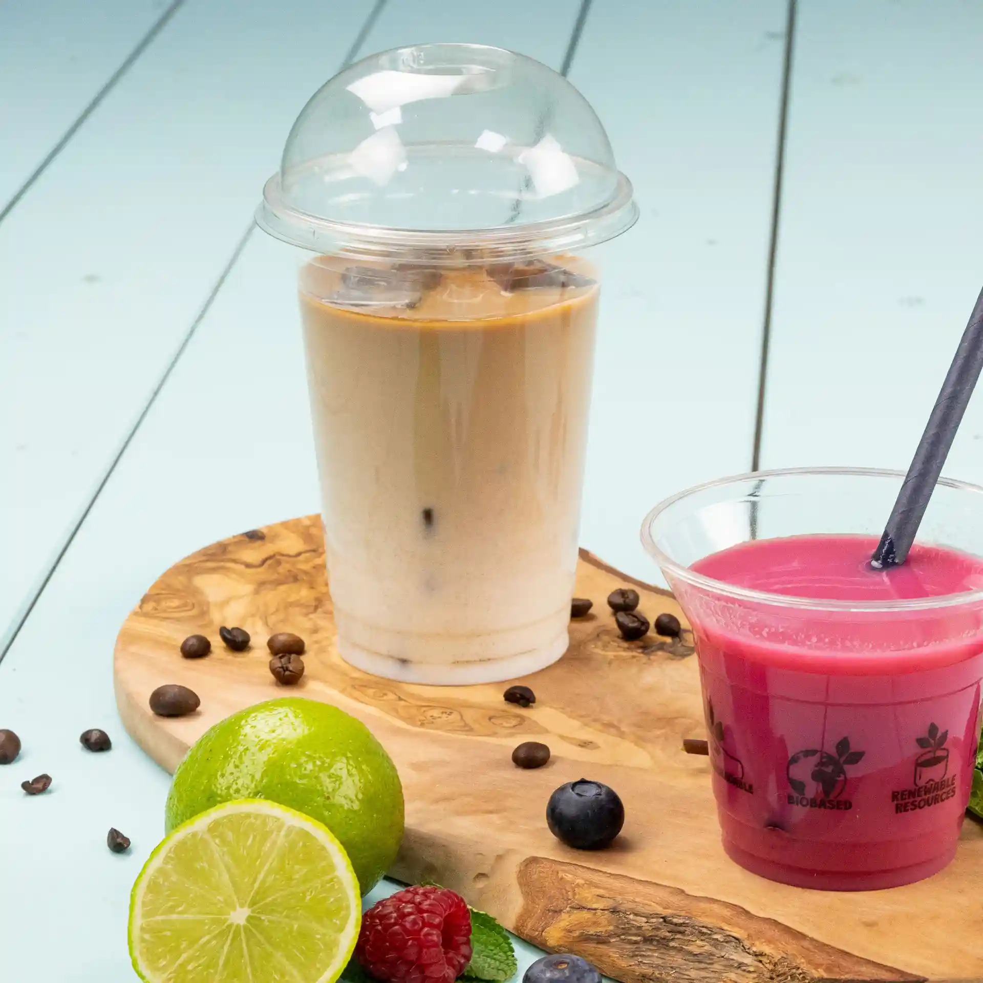 Image d'un gobelet en plastique transparent de 16 oz rempli de café glacé, à côté d'une boisson rose dans un gobelet transparent sur une planche en bois avec des fruits.