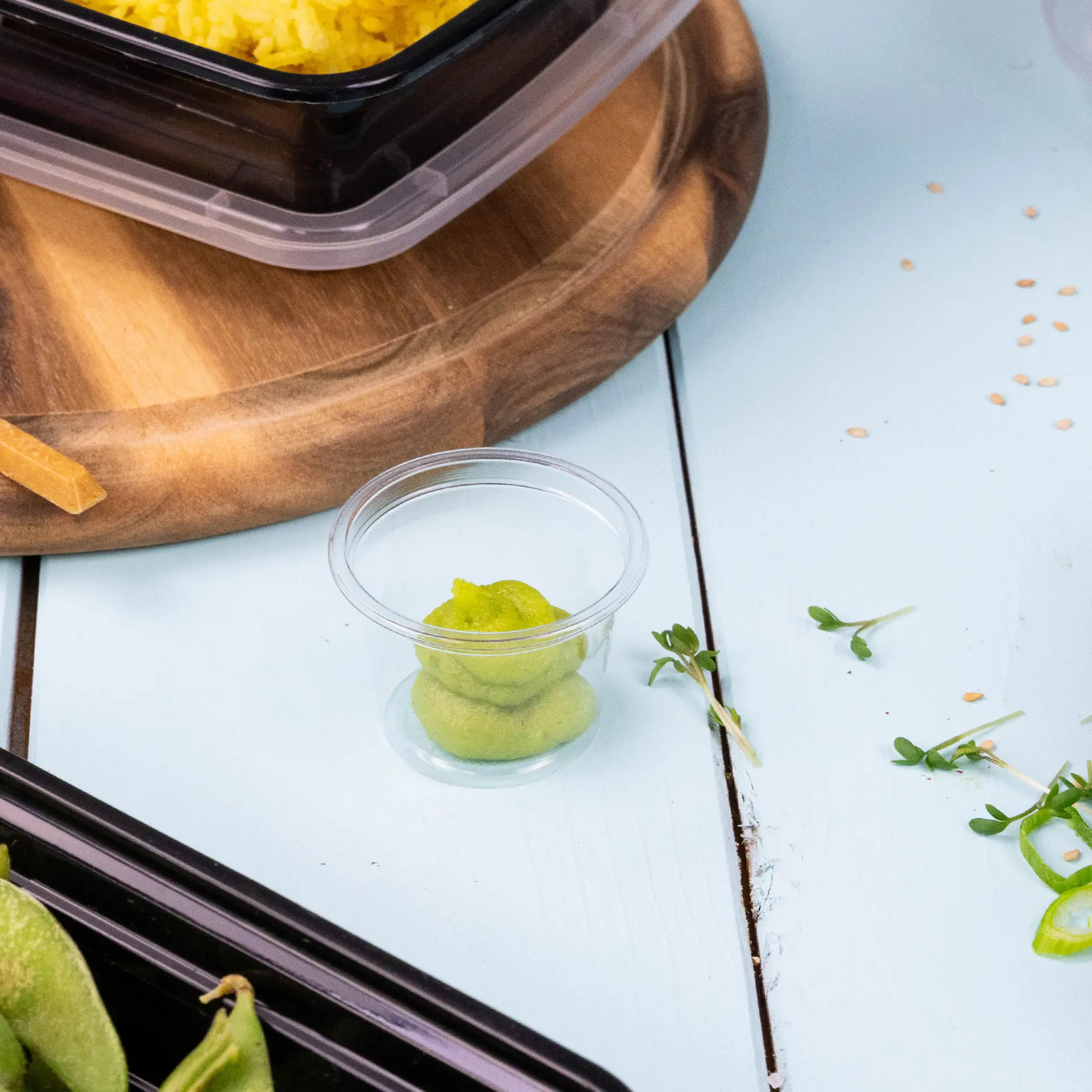 Un gobelet en plastique transparent avec du wasabi vert à l'intérieur, posé sur une surface en bois bleu clair près d'emballages alimentaires et de garnitures.