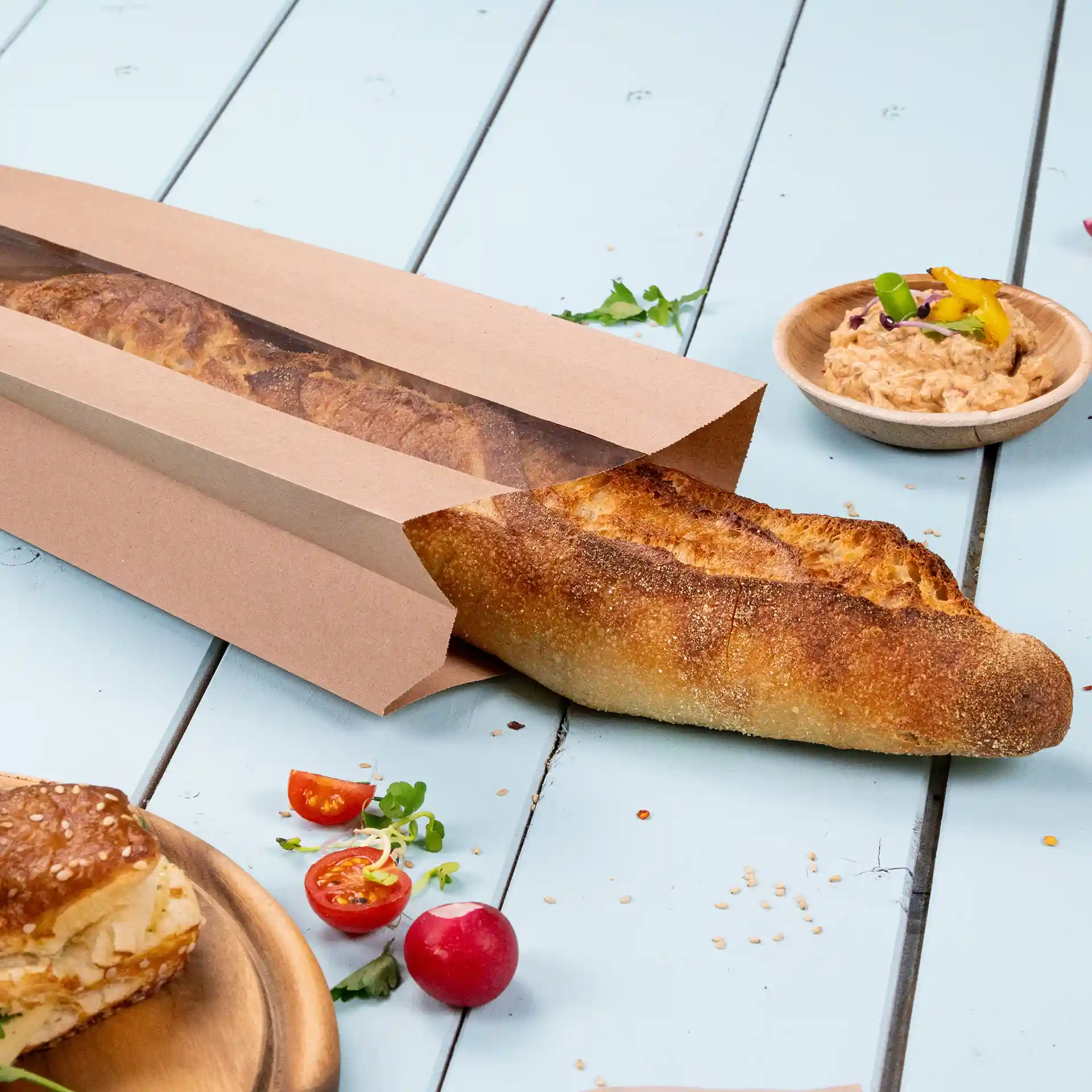 Sac à pain en papier brun plat avec une fenêtre, contenant une baguette, placé sur une surface bleu clair avec des tomates et une sauce.