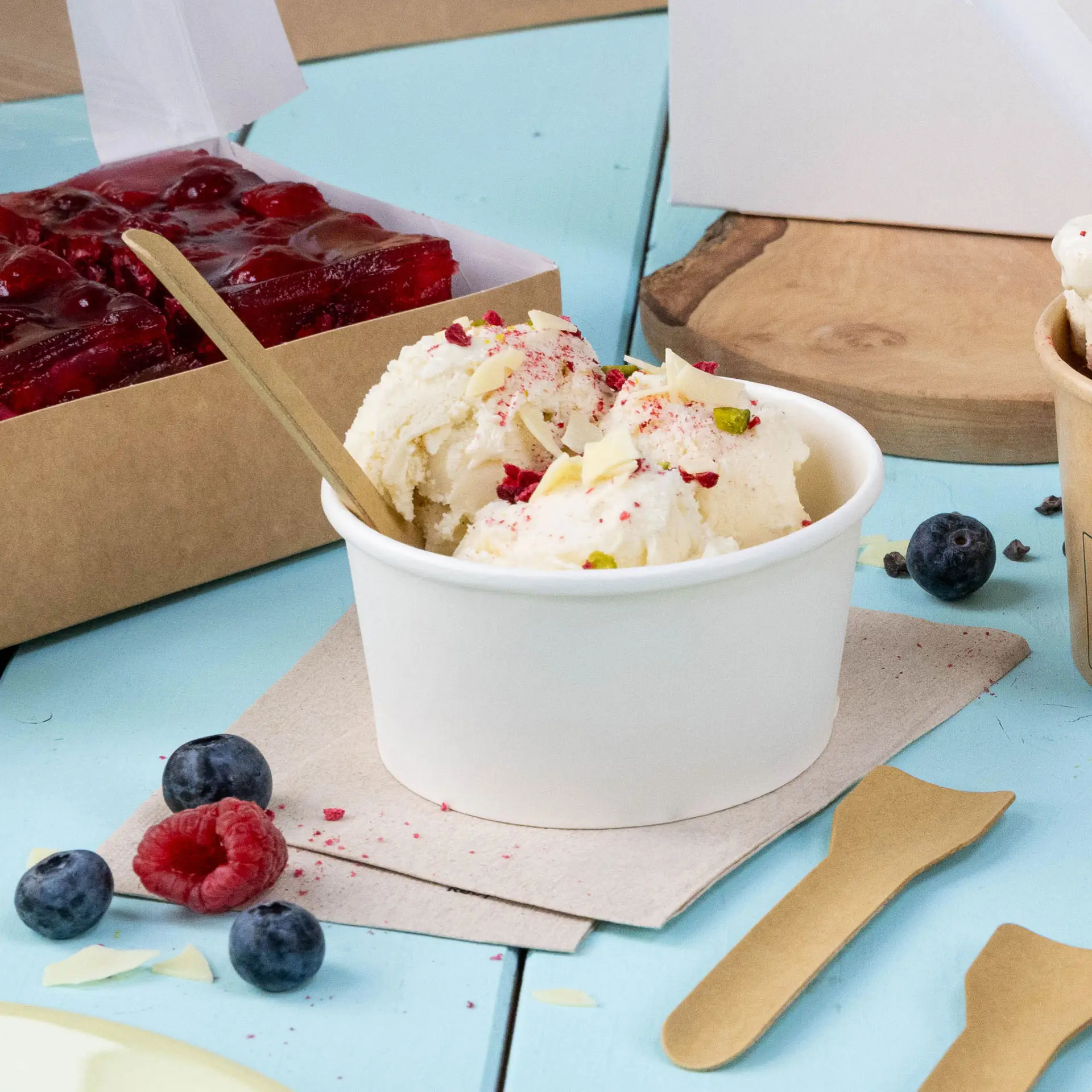 Un verre de glace surmonté de fruits et de noix, cuillère en bois et baies fraîches sur une surface bleue avec une serviette.