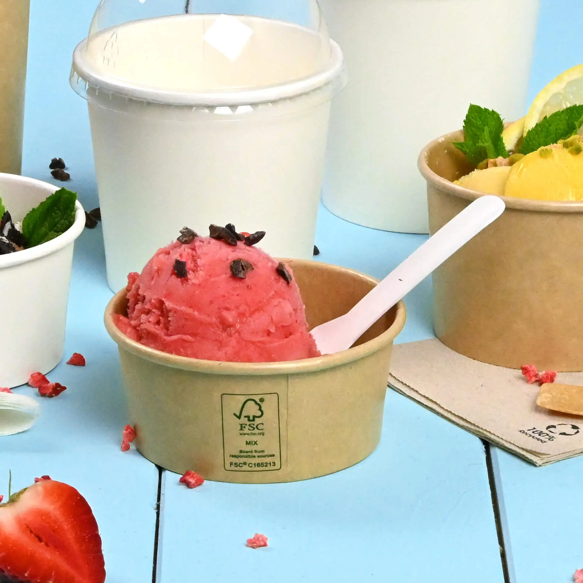 Un gobelet en papier brun avec de la glace rose, garnie de pépites de chocolat ; d'autres gobelets de glace sont partiellement visibles en arrière-plan.