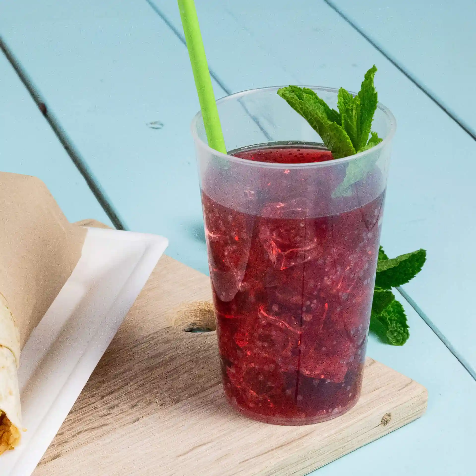 Gobelet réutilisable transparent rempli d'une boisson rouge et de glace, avec une paille verte et des feuilles de menthe, posé sur une surface en bois.