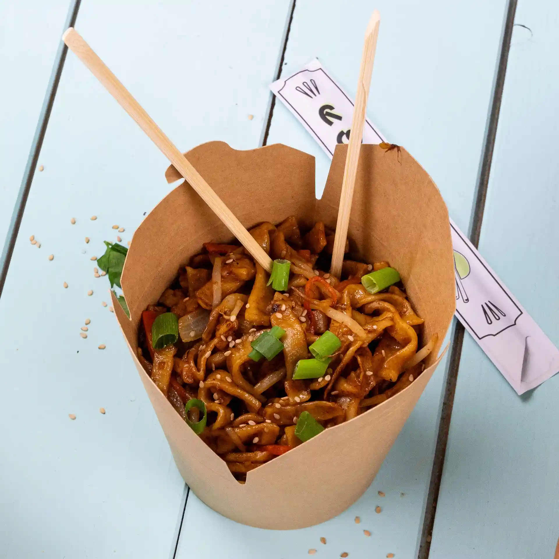Boîte à nouilles en carton brun remplie de nouilles sautées, garnie d'oignons verts et de graines de sésame, avec des baguettes insérées.