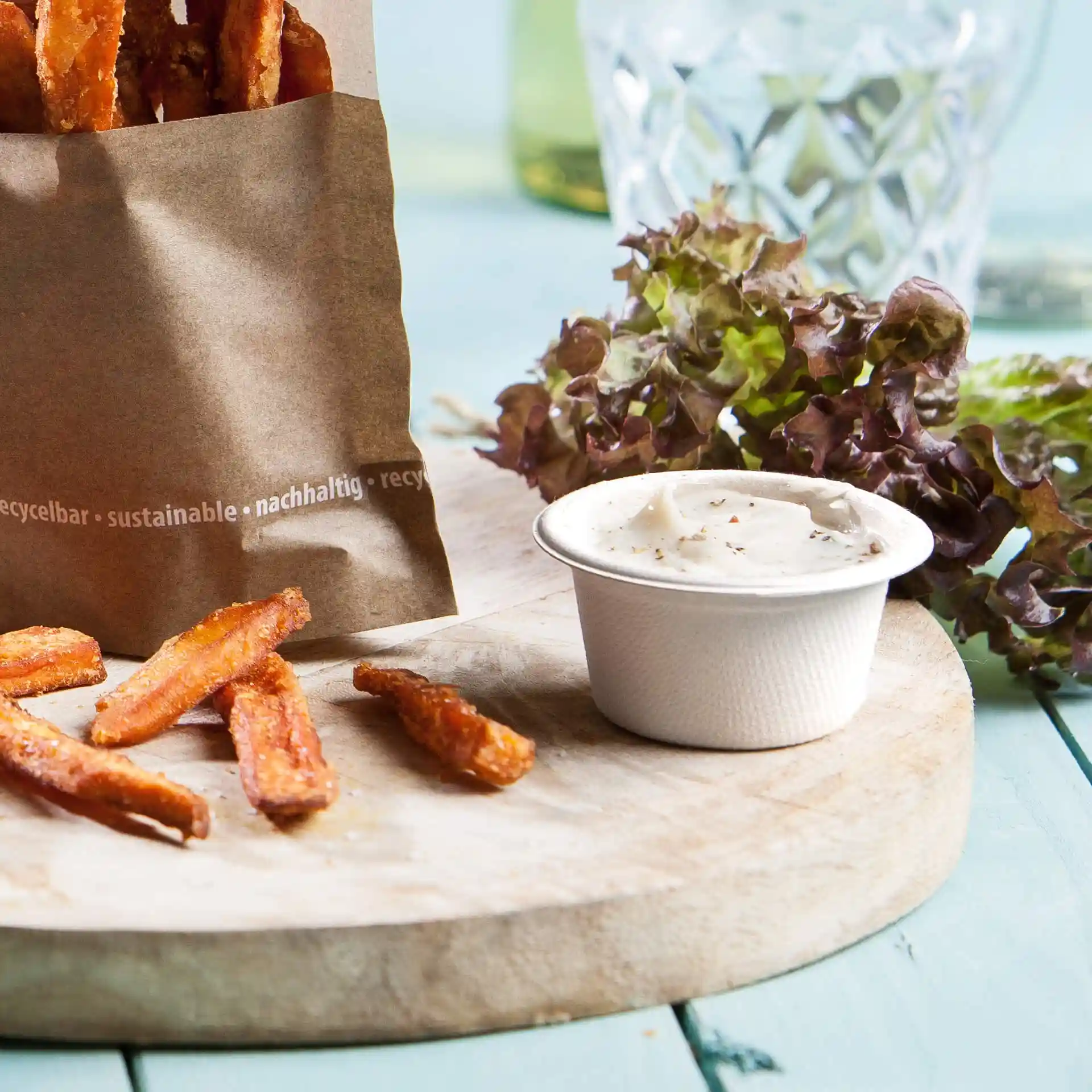 Frites de patate douce dans un sac en papier, avec un petit pot de sauce et de la laitue fraîche sur une planche en bois.
